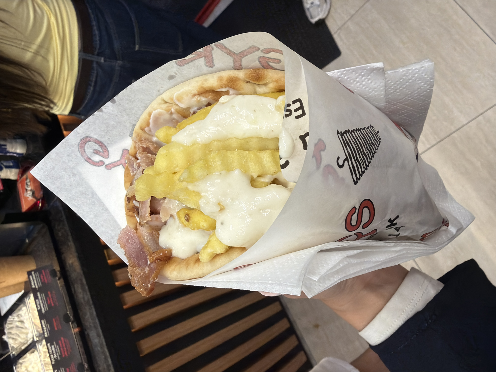A person holds a gyro sandwich wrapped in paper. The gyro is filled with meat, french fries, and a creamy white sauce. The paper wrap has red and black text and graphics, including what appears to be an umbrella illustration. In the background, another person is wearing a yellow shirt and blue jeans, and dark-colored clothing can be seen. The setting appears to be indoors, possibly a food establishment, with a tiled floor visible in the upper right corner. The overall impression is casual, likely a casual meal or snack. The visible text on the wrapper is difficult to read clearly, but some letters like "EX", "ES", and "S" are discernible, along with some Cyrillic characters. The lighting suggests it is daytime.
