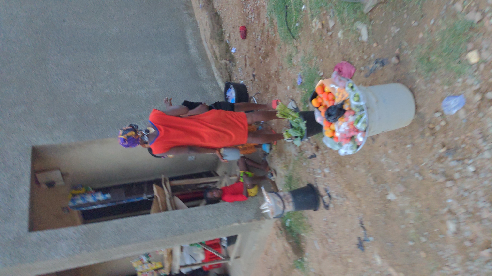 A slightly blurred outdoor image captures a woman dressed in an orange tunic with blue trim and a patterned headwrap. She stands on an unpaved ground area adjacent to a building with a concrete exterior. The woman appears to be selling items from a bucket that is overflowing with tomatoes, some leafy green vegetables, and other wrapped items that are hard to identify. A small girl is lying nearby and a young child is standing to the side. The location is characterized by a dusty, somewhat cluttered environment, suggestive of a roadside setting, and is in Ado, Nigeria.