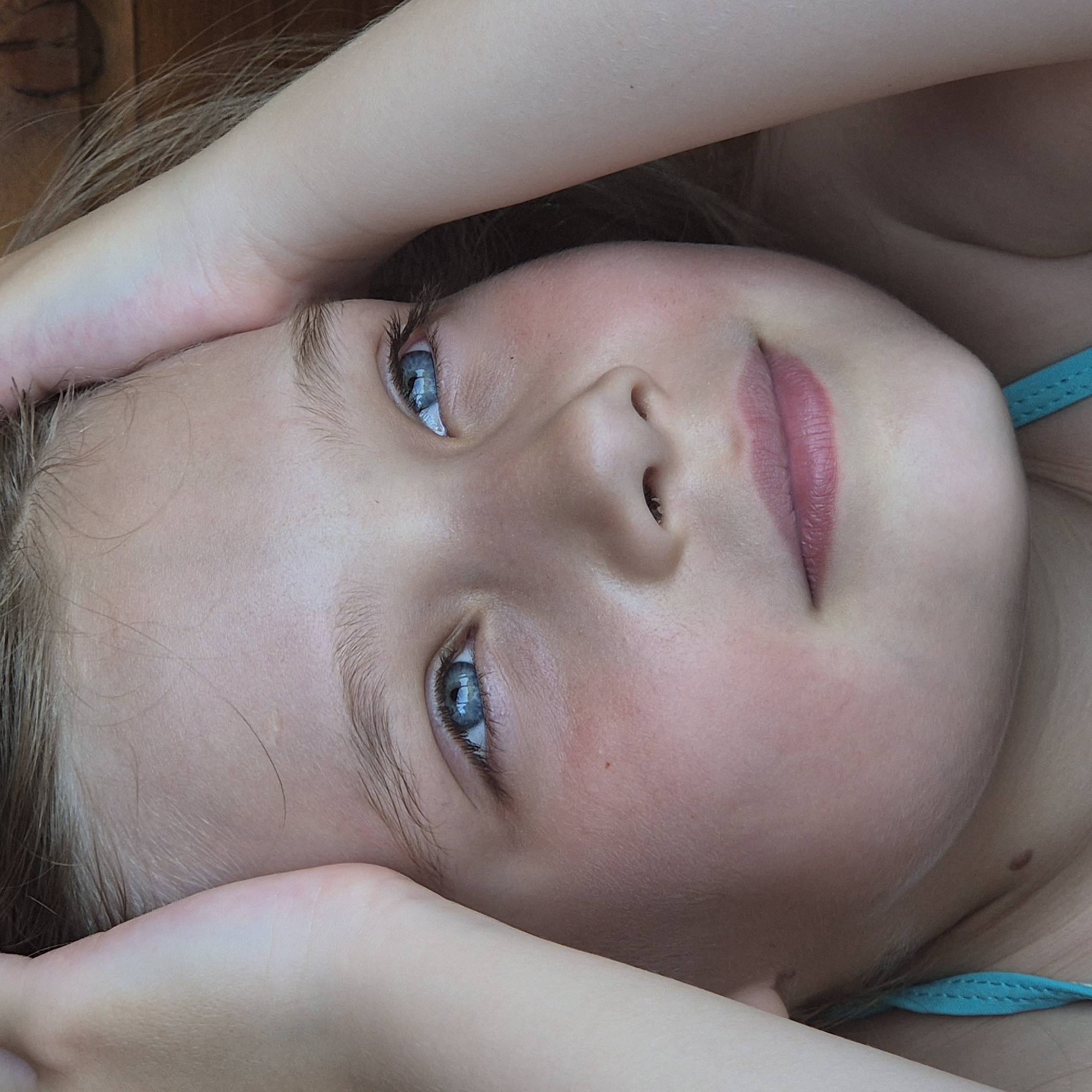 The image portrays a young Caucasian individual, likely female and appearing to be a child or pre-adolescent, captured in a close-up, head-and-shoulders composition. The subject's hands are raised, with forearms positioned along the sides of the head and hands gently resting near the temples. The gaze is directed upwards and slightly to the viewer's right, indicating a pensive or observant expression.
The individual has fair skin with natural pinkish undertones, particularly on the cheeks. Light brown or blonde hair frames the face, with fine strands visible across the forehead and around the ears. The eyebrows are light brown and moderately full. Prominent blue eyes exhibit visible pupils and distinct catchlights from an ambient light source, reflecting light on the irises. A small, dark brown mole is located below the viewer's left eye, and another is visible on the lower neck/upper chest area on the viewer's left side. The nose is small and the lips are closed, displaying a natural pink hue.
A turquoise-colored strap, consistent with a swimsuit or tank top, is visible on the lower left side of the frame. The background is softly blurred, providing a shallow depth of field. A dark, textured surface, possibly wood, is discernible behind the subject's right side, while the left background is an indistinct, darker blur. The scene is located in Новостройка, Russia.