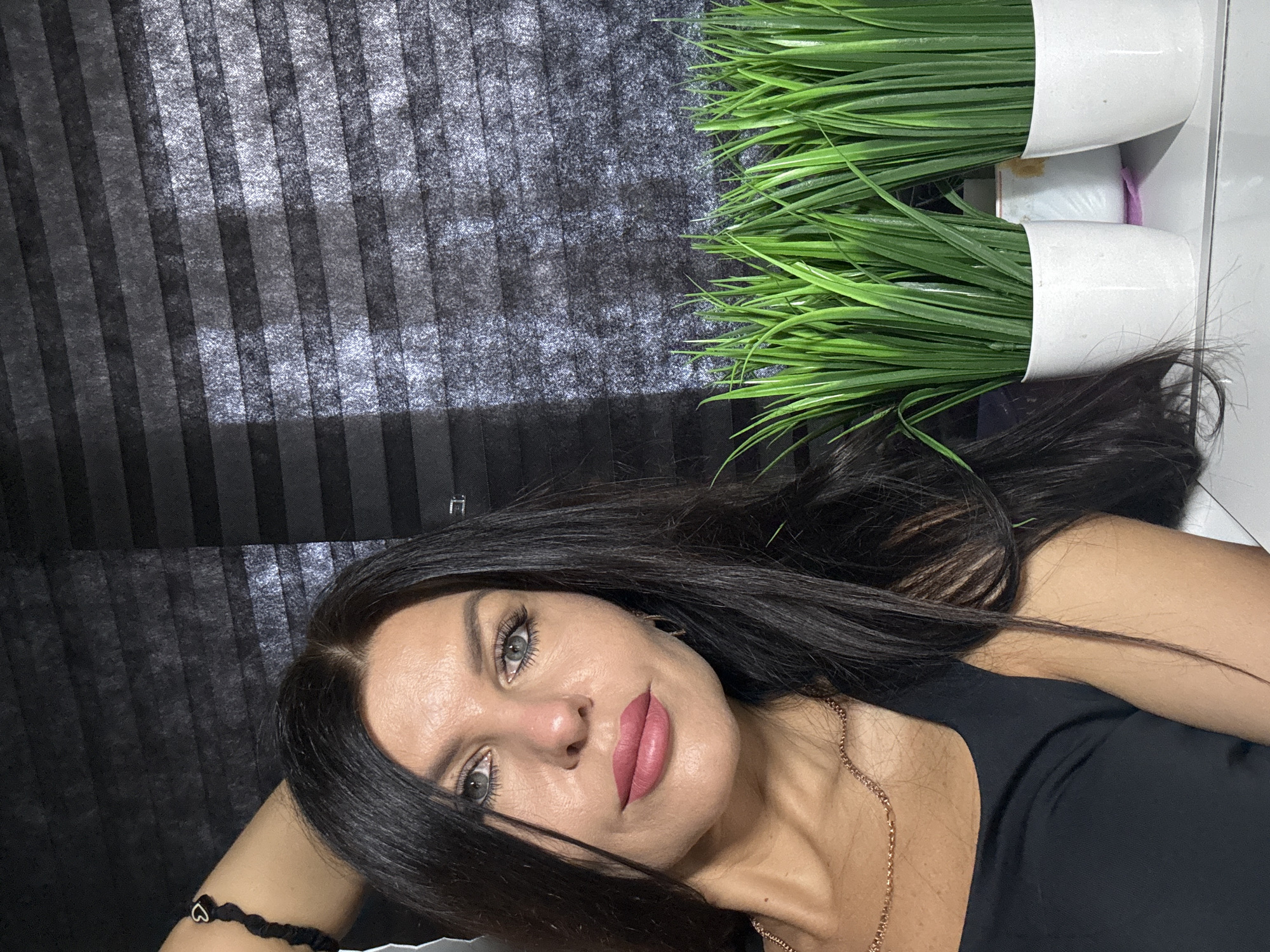 A close-up, diagonally oriented portrait features a woman posing indoors. Her head is in the lower left of the frame, resting on her left hand, while her body extends towards the upper right. She has long, dark hair that cascades around her shoulders, and striking light-colored eyes. Her face is made up with pink lipstick and visible mascara. She wears a black top, a delicate golden chain necklace, and a black bracelet adorned with a small silver heart charm on her wrist.
The background consists of dark, textured, striped blinds or curtains in the upper left portion of the image. In the upper right, two artificial green plants in white cylindrical pots are visible, suggesting a decorative arrangement on a shelf or surface behind her. The overall lighting is soft, indicative of an indoor setting, and the woman maintains a neutral, direct gaze towards the camera. No text is visible in the image. The scene is set in Chaikovsky, Russia.