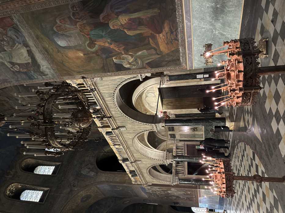 The image depicts the interior of a large Orthodox cathedral, identified as the Alexander Nevsky Cathedral in Sofia, Bulgaria. The perspective is from the ground floor, looking towards an arched arcade and a prominent side wall.
In the immediate foreground, two ornate, copper-toned metal candle stands are visible. The stand on the right is more fully in view, circular and multi-tiered, holding approximately twenty slender white candles, all actively burning and casting warm light. The stand on the left is partially cropped. The floor beneath is a checkered pattern of light and dark grey or black tiles.
The midground features significant architectural elements. A series of three dark, polished columns support arches with intricately carved spandrels. Above these arches, a balustraded gallery or balcony with lighter, possibly marble, stonework extends across the scene. Beyond this arcade, further interior spaces are visible, including another dark entrance or doorway.
Approximately four individuals are present within the visible area. To the left of the central column grouping, three men in dark coats or jackets are gathered, seemingly conversing. Further to the right, a lone individual in dark clothing walks near a column and another doorway.
The right wall is dominated by a large, colorful religious mural depicting multiple biblical figures. A central figure, resembling Jesus, is shown interacting with several individuals, some of whom are kneeling. The mural features an earthy color palette with prominent blues, reds, and whites. Below the mural, the wall is paneled with green-marbled stone.
Overhead, a massive, elaborate metal chandelier with multiple arms, each holding unlit candle-shaped bulbs, hangs centrally from the high, vaulted ceiling. The ceiling itself is adorned with dark, faded murals. In the upper-left, high arched windows with grid patterns allow ambient light into the space. The overall lighting is dim, augmented by the warm glow of the lit candles.