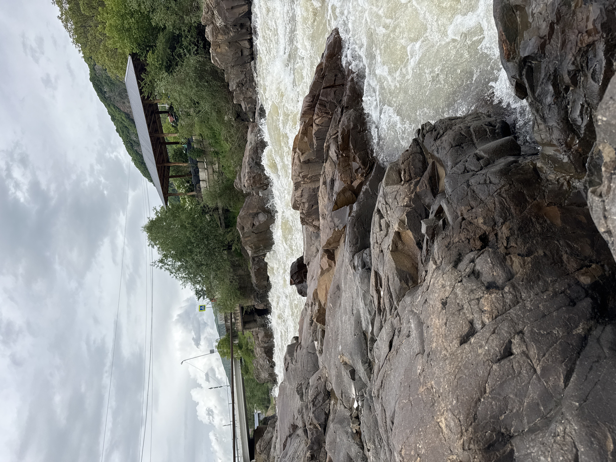 The image depicts a rushing river flowing over rocks, with a pavilion visible on the left bank. The sky is overcast. The location is Каменномостский, Russia. A bridge and street lamps can be seen in the distance.