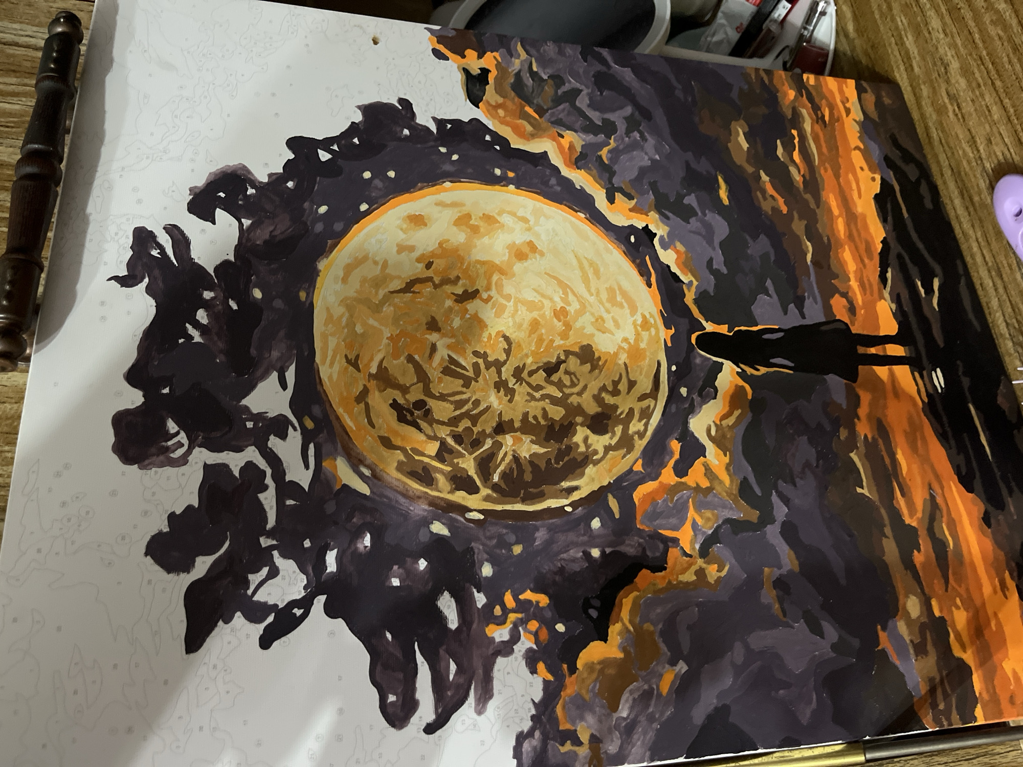 The image is a partially completed paint-by-numbers project, depicting a scene with a large, yellow moon-like object in the center. To the left of the moon is dark, amorphous shapes resembling trees or clouds. The right side of the image shows a silhouetted figure or figures standing against a backdrop of orange and dark brown shapes, possibly representing trees or other structures. The canvas is white with numbered outlines of the areas to be painted. The painting is set on a wooden surface. There is no indication of the city of Chaikovsky, Russia.