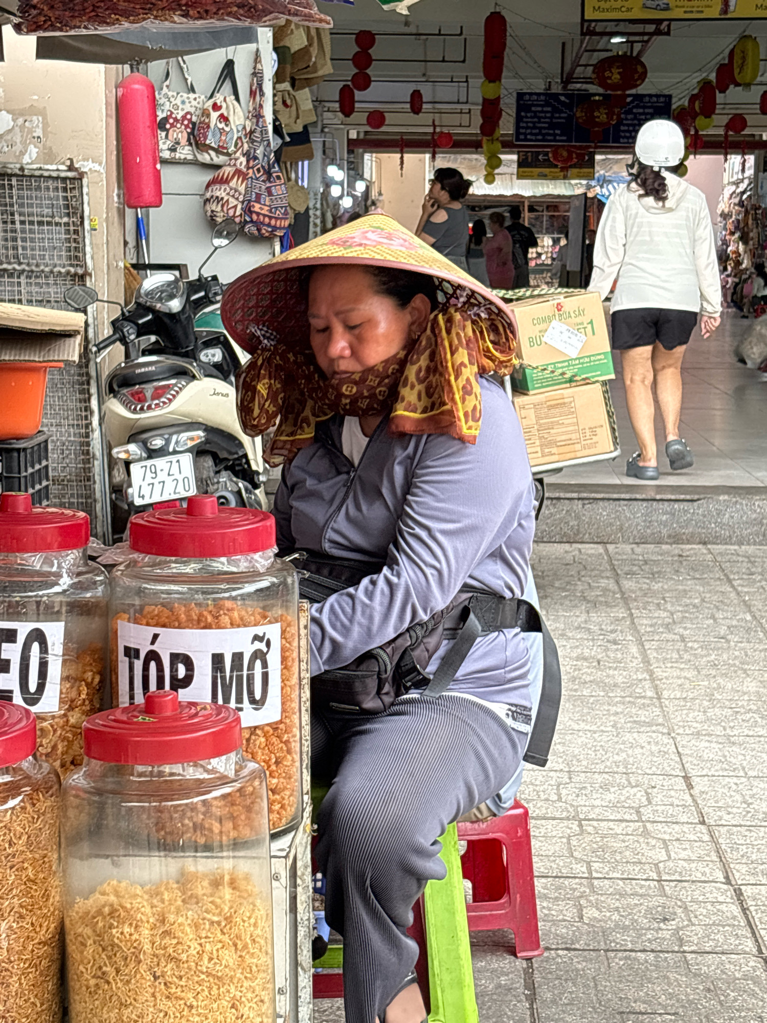 This is a medium shot of a woman sitting on a small red stool in front of a street stall in Nha Trang, Vietnam. She is wearing a conical hat and a patterned scarf around her neck. The stall displays several large glass jars filled with what appear to be dried food items, possibly noodles or snacks. Behind her, a motorcycle is parked and a variety of bags are hanging from the stall. In the background, other people are walking through a busy market area. The scene appears to be in an outdoor market environment typical of urban street commerce in Vietnam.