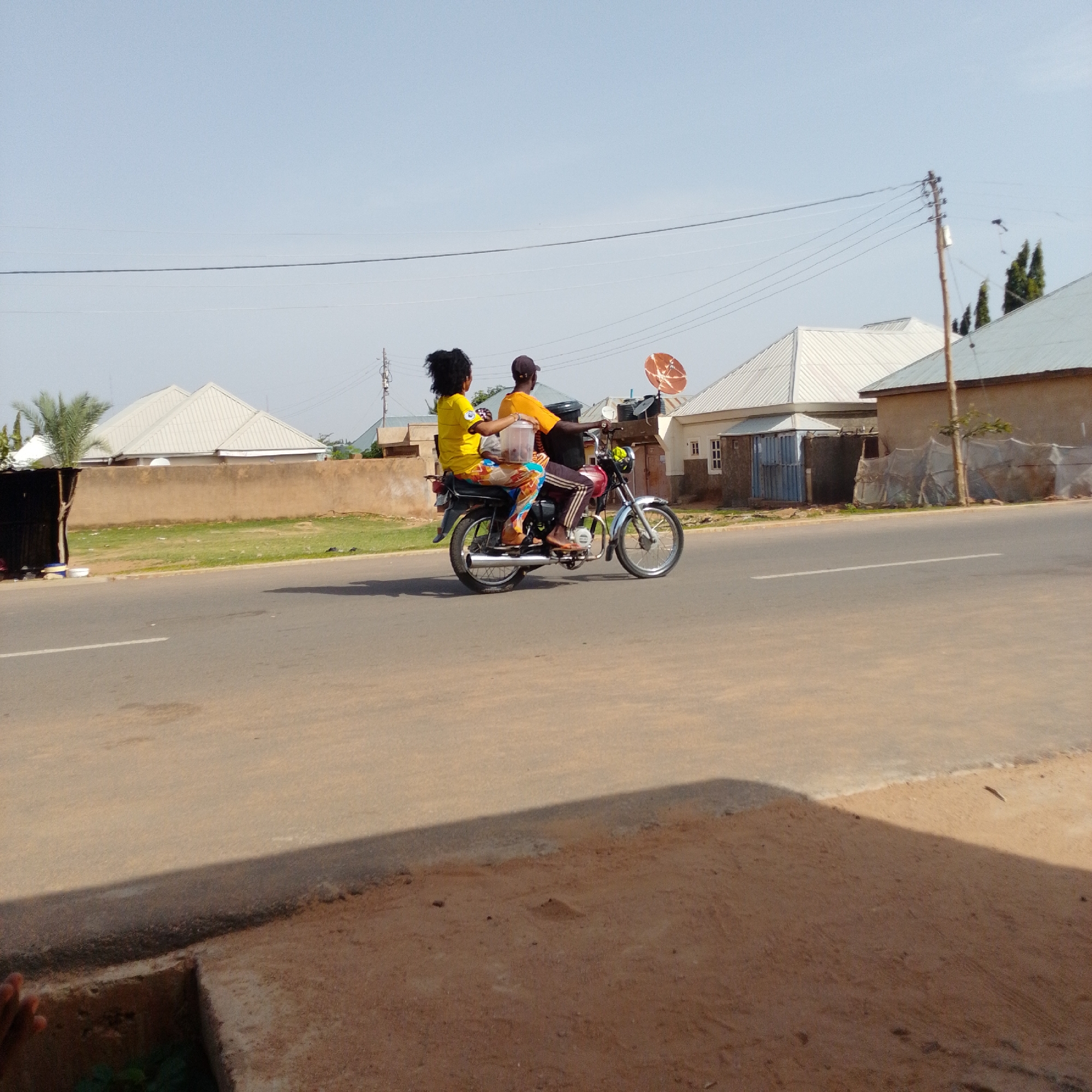 In Gombe, Nigeria, a motorcycle carrying two people rides on a paved road. The driver, wearing a dark cap and an orange shirt, operates the vehicle. The passenger, a woman with dark, curly hair, is dressed in a yellow shirt and patterned trousers, and she holds a clear plastic container with a dark lid. The motorcycle is a dark red color with chrome accents. Behind them, buildings with white, sloped roofs are visible, along with a beige wall and some greenery. Telephone poles and wires stretch across the pale blue sky, and a satellite dish is mounted on one of the buildings. The road surface is asphalt, with a white line marking the lane. The foreground shows a patch of reddish-brown soil next to the road.