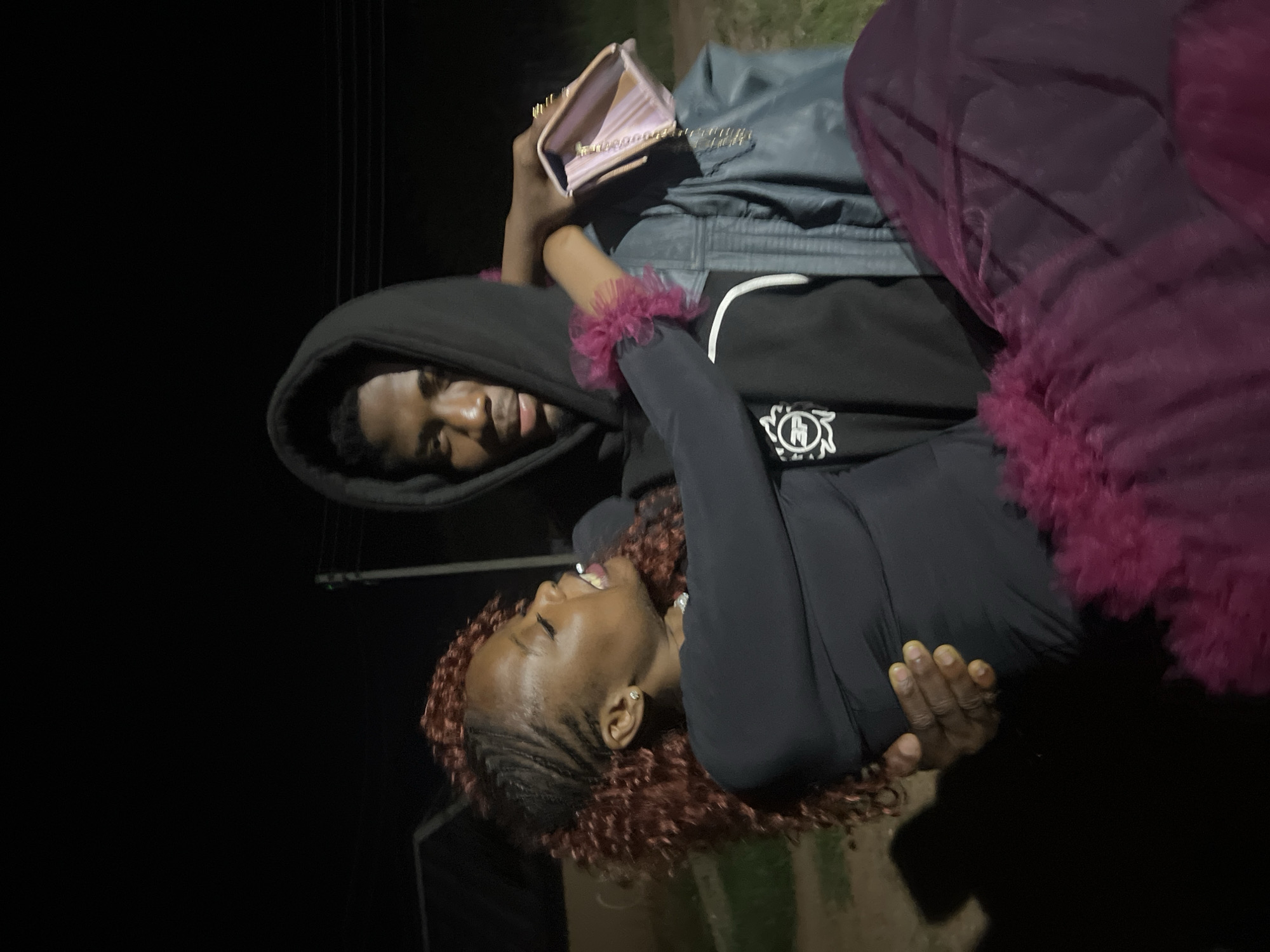 The image depicts two young adults in Durumi, Nigeria, at night. The man, wearing a black hoodie and a denim jacket, is carrying the woman. The woman, with reddish curly hair styled with braids, is wearing a black long-sleeved top and a voluminous, dark pink skirt. She is leaning towards the man, with her eyes closed and mouth open, appearing to be in a playful or intimate moment. She is holding a light pink clutch purse in her right hand. The background is dark, indicating nighttime, with some faint lights and structures suggesting an outdoor, possibly rural or semi-urban, setting.
