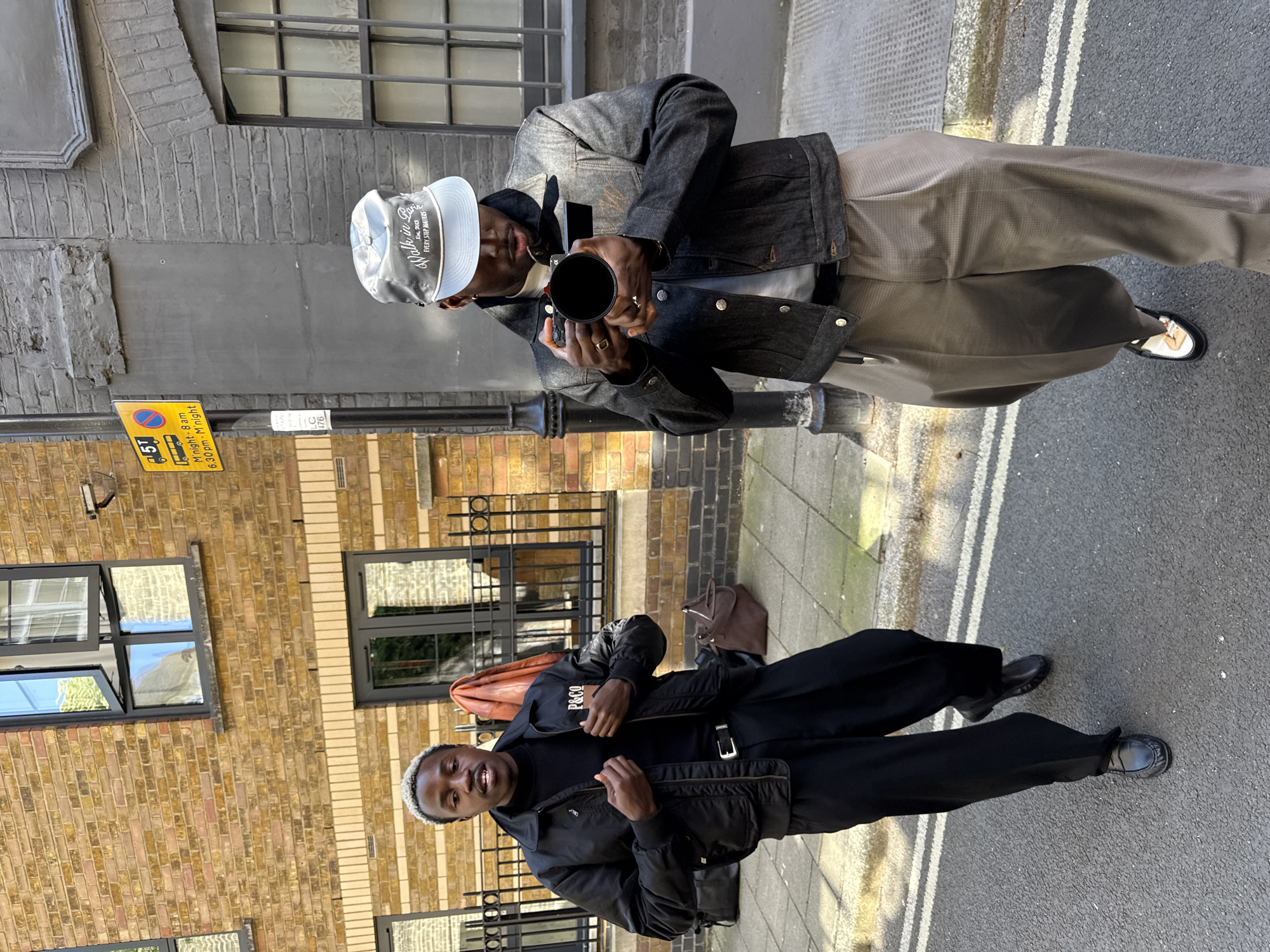 The image shows two men standing on a street in London, United Kingdom. The man in the bottom frame is wearing a black jacket with "P&CO" visible on the inner label, a black turtleneck, black trousers with a belt, and black shoes. He is smiling slightly and adjusting his jacket. Behind him is a brick building with dark-framed windows. A brown leather bag is on the ground. The other man in the top frame wears a denim jacket, light-colored trousers with a subtle grid pattern, and two-tone shoes. He is holding a black cylindrical object (possibly a camera lens) up to his face, partially obscured by his hands. He also wears a baseball cap with text that reads "Walk in Paris, Est. 2003, EVERY STEP MATTERS." Behind him is a brick building with gray painted upper sections. The street is paved with concrete slabs and has painted white lines marking lanes. A yellow parking sign with black text and graphics is also visible on the left side of the frame. The street appears to be a relatively narrow lane or alleyway.