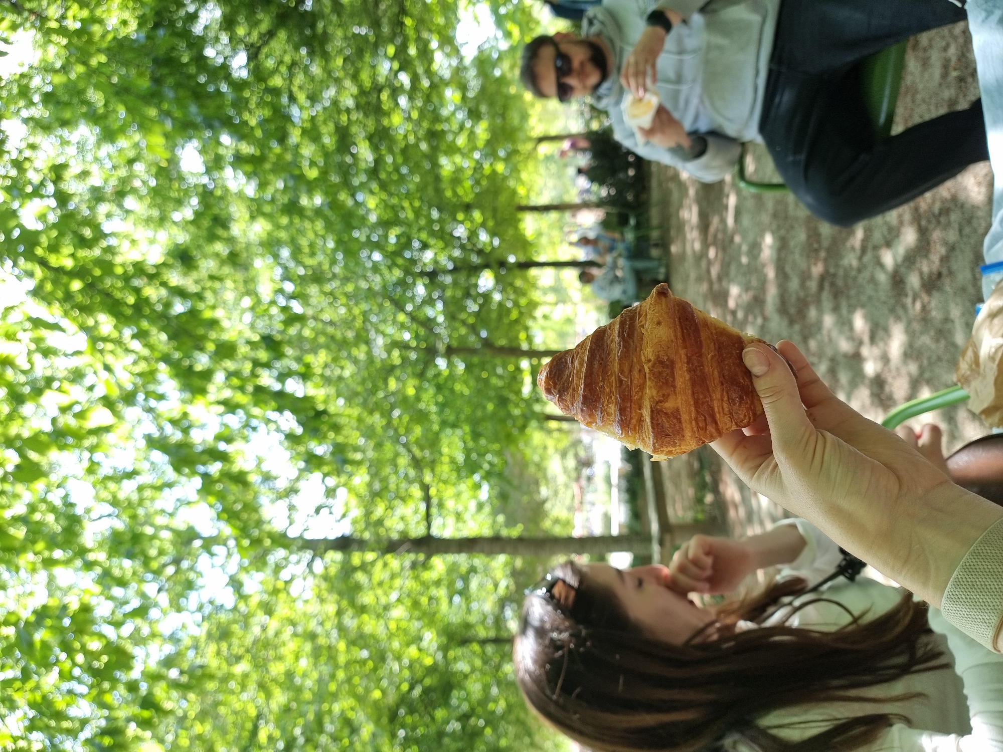 The image captures an outdoor scene in what appears to be a park or garden, likely in Paris, France, given the presence of a croissant. The photo is taken at a tilted angle. A woman is in the foreground, holding a croissant in her right hand, with her mouth open as if she's about to take a bite. She has long, brown hair and wears sunglasses on her head, with a white shirt or blouse.
In the background, a man is sitting on a green metal park chair, wearing sunglasses and eating something from his hands. He wears a gray hoodie, dark jeans, and a watch on his left wrist. Other people are sitting on benches further back, blurred into the scene. The environment includes many green trees, some with dense foliage that creates a blurred, bokeh-like effect in the background. The ground appears to be covered with gravel or sand. The overall scene suggests a casual, relaxed atmosphere in a public park.