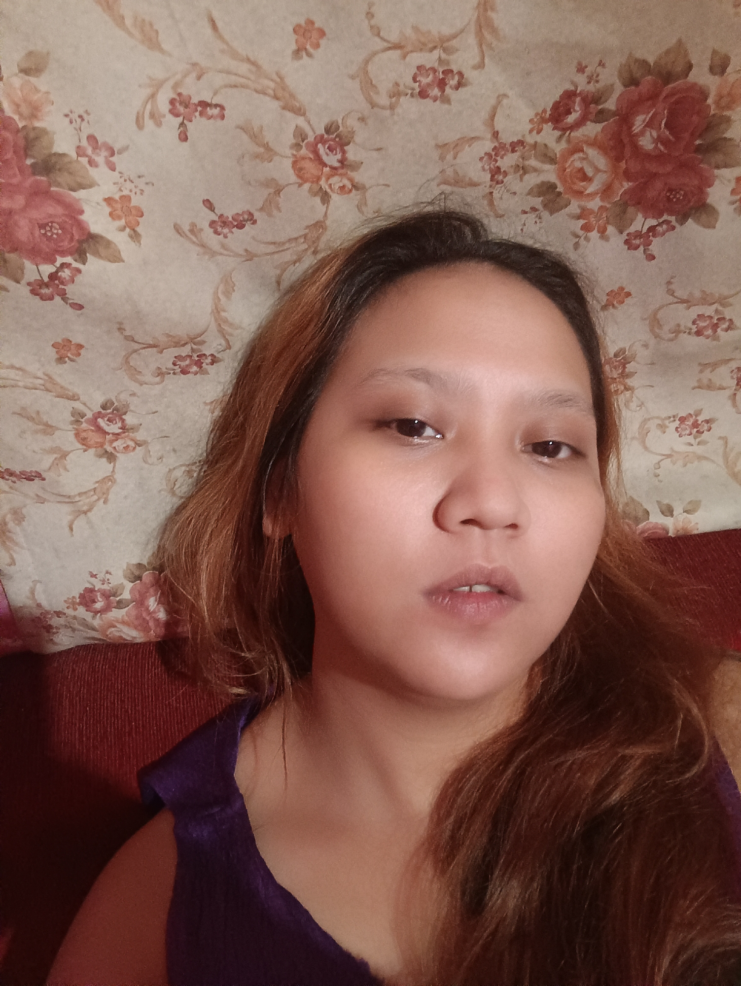 This is a close-up, eye-level, selfie-style photograph of a young woman. She is positioned in the center of the frame, with her face taking up a significant portion of the image. The background consists of floral wallpaper with a pattern of pink and red roses and brown floral accents on a light beige background. She has medium-toned skin and shoulder-length, wavy, reddish-brown hair that frames her face. Her eyes are dark brown and her eyebrows are light brown. Her mouth is slightly open, revealing her teeth. She is wearing a dark purple top. The lighting suggests an indoor setting, possibly during the day or evening, with no strong shadows indicating direct sunlight. The overall mood appears relaxed or contemplative. There is no visible text in the image. The context provided places the scene in Depok, Indonesia.