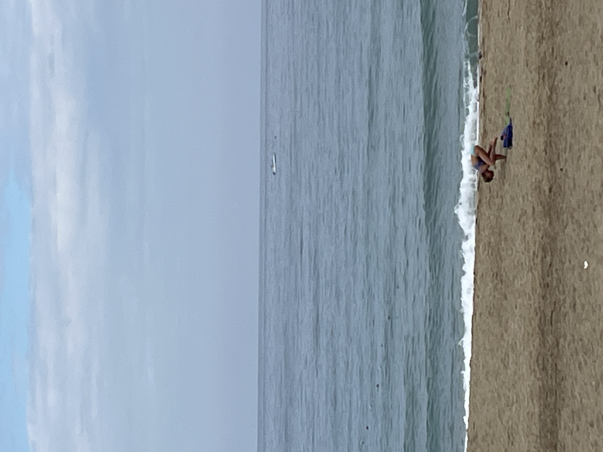 The image depicts a beach scene. A person is standing near the water's edge. To the left, there is a calm body of water and the sky. The location is likely the city of Alanya, Turkey.