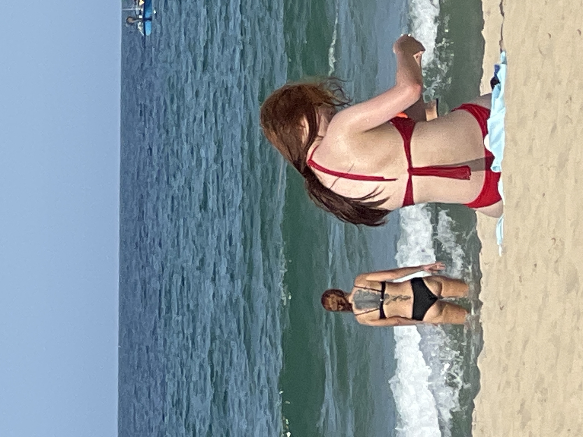 The image captures a scene on a beach in Da Nang, Vietnam. Two women are present. One woman, with long reddish-brown hair, is seated on the sand in the foreground, facing away from the camera and towards the ocean. She is wearing a red bikini. Her arms are positioned in front of her, and she appears to be engaged in an activity with her hands, though it is not entirely clear. The other woman, positioned further back and in the water, also faces away from the camera. She has dark hair tied up and is wearing a black bikini. A large, detailed tattoo covers her back. Gentle waves are breaking on the shore, creating white foam. The water is a deep blue-green, and the sky above is a pale blue. In the distance, a small blue boat is visible on the horizon. The sand in the foreground is light brown and appears dry. A light blue towel is laid out on the sand near the seated woman.