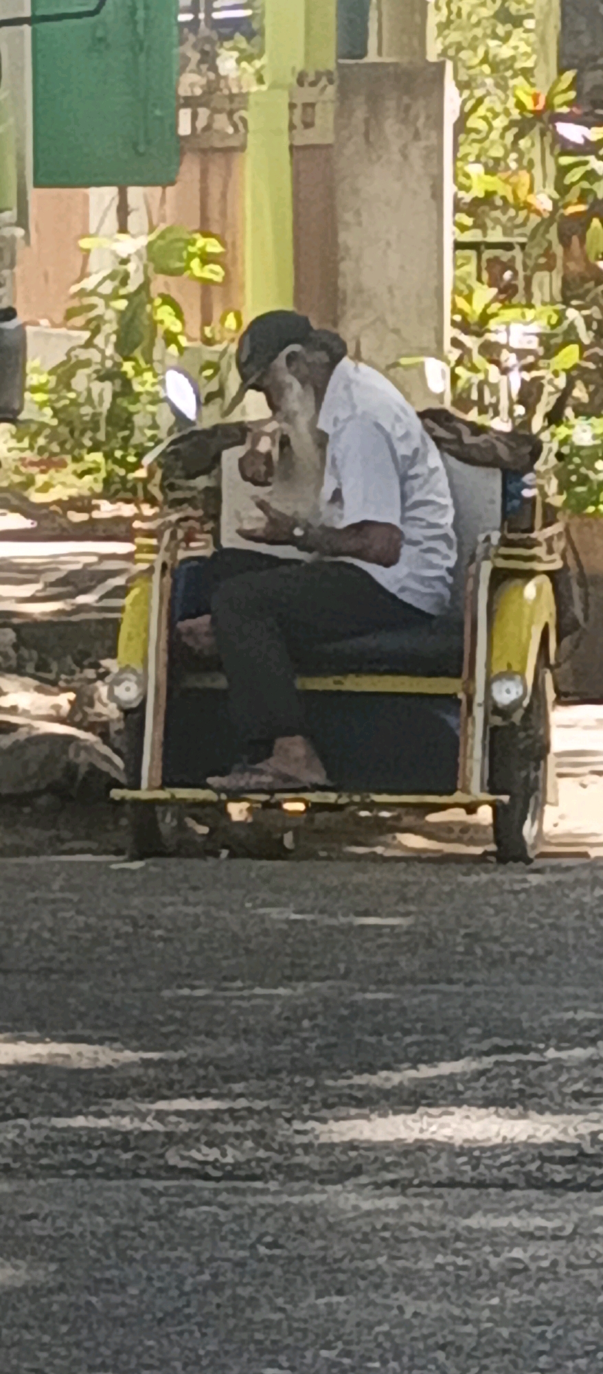 The image depicts an elderly man with a long white beard, wearing a white short-sleeved shirt, dark pants, and a dark baseball cap. He is seated in the passenger compartment of a yellow auto-rickshaw, also known as an "ojek" or "becak motor" in Indonesia. The man is holding a small object, possibly a cigarette or a small snack, in his right hand and appears to be bringing it towards his mouth. His left hand is held open, gesturing slightly. The auto-rickshaw has a metal frame with yellow body panels and black upholstery in the passenger area. The wheels are visible, and there are lights on the front and side of the vehicle. The background shows a street scene with vegetation, including green bushes and flowering plants, and a building with some architectural details, possibly a wall or a column. The lighting suggests it is daytime, with sunlight casting shadows. Based on the presence of an auto-rickshaw and the typical urban environment, this scene is likely from Malang, Indonesia.