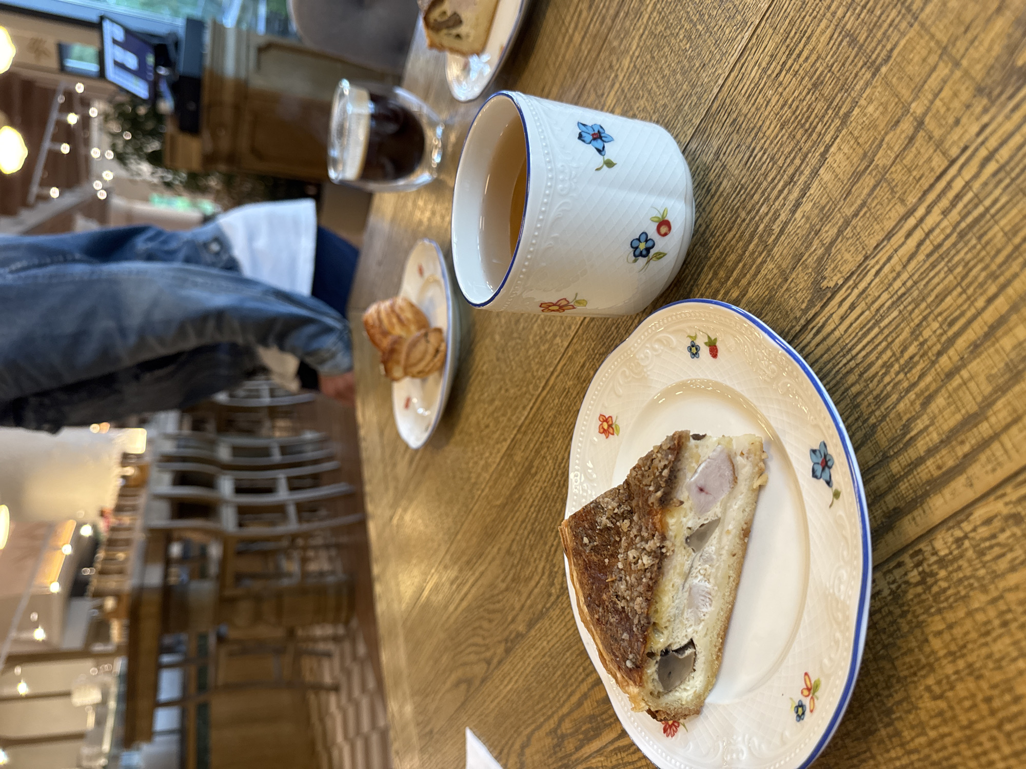 The image shows a table setting with a plate of quiche, a cup of tea, a glass of dark liquid (likely coffee or tea), and a small plate of pastries. All items are on patterned china. The table is made of wood, and in the background, a person's legs in denim jeans are visible, along with a glimpse of a window and lighting fixtures. The location is potentially a café or restaurant. The image doesn't provide enough detail to confidently confirm the location as Izhevsk, Russia. More specific information about the interior design or other contextual clues would be needed.