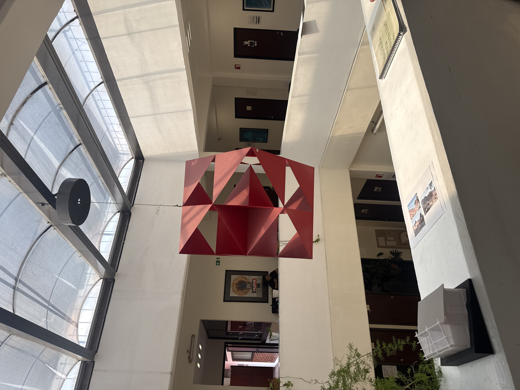 The image captures the interior of a building, likely a multi-story structure, located in San Andrés Cholula, Mexico. A prominent feature is a large, three-dimensional, red, geometric sculpture suspended in the open space between floors. The building's architecture incorporates white walls, and beams and a skylight that allows natural light to illuminate the space. Several floors can be seen receding into the distance, each with different posters, signage, and plants. A person is partially visible on one of the floors near the sculpture. Other wall decorations or displays are visible on each level, with more greenery near the bottom. The presence of potted plants suggests an attempt to bring a natural element into the indoor environment. The skylight area is covered in white shading of some type.