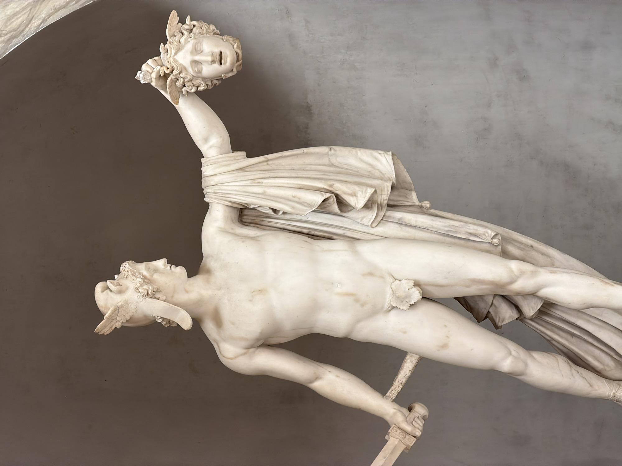 A full-length, polished white marble statue of Perseus with the Head of Medusa stands against a plain, light brown-grey wall, illuminated from above, within the Vatican Museums in Città del Vaticano, Vatican City. The male figure, Perseus, is depicted with a muscular, nude physique, his genitals covered by a carved fig leaf. He wears a winged helmet on his head and looks towards his right. His right hand holds a sword, its hilt visible, with the blade resting along his right leg. His left arm is extended upwards and forward, holding the severed head of Medusa by its hair, which is represented by snakes. A draped cloak hangs over Perseus's left arm and shoulder, falling to his mid-thigh. The statue rests on an unseen base. The background wall is unadorned, providing a neutral backdrop.