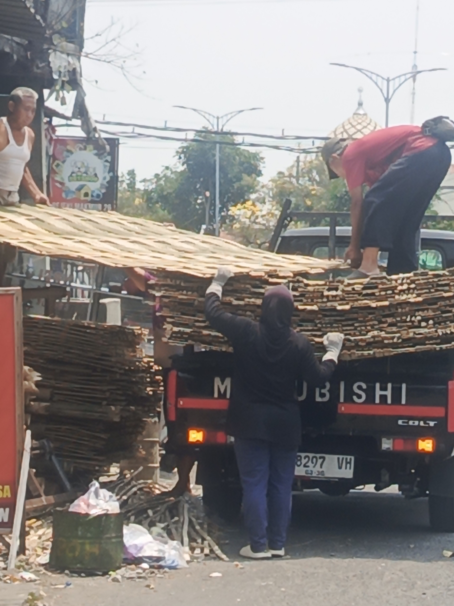 The image depicts a street scene in Surabaya, Indonesia, where individuals are engaged in loading or unloading a significant quantity of wooden planks onto a truck.
In the foreground, a person wearing a dark hooded jacket and blue pants, with gloves on their hands, is actively involved in the task, positioned on the truck bed. Another person in a red shirt and black pants, wearing a cap, is also seen near the top of the stacked planks. A third individual, an elderly man wearing a white tank top, is visible on the left side, leaning over the planks.
The primary subject is the truck, which appears to be a Mitsubishi Colt, identified by the visible "MITSUBISHI" and "COLT" lettering on its tailgate. The truck is laden with stacks of thin, light-colored wooden planks, forming a substantial pile. Some of these planks are also visible in stacks next to the truck and on the ground.
The environment suggests an outdoor setting, likely a roadside in Surabaya. Evidence of urban surroundings includes the presence of a street, a paved surface, and what appear to be buildings or stalls in the background. There is also visible debris, including a green barrel and plastic bags, scattered on the ground, indicating a somewhat unkempt area. Overhead, power lines and some tree branches are visible against a bright sky. The overall impression is a bustling, perhaps commercial, area of Surabaya where manual labor is being utilized for the transport of goods.
