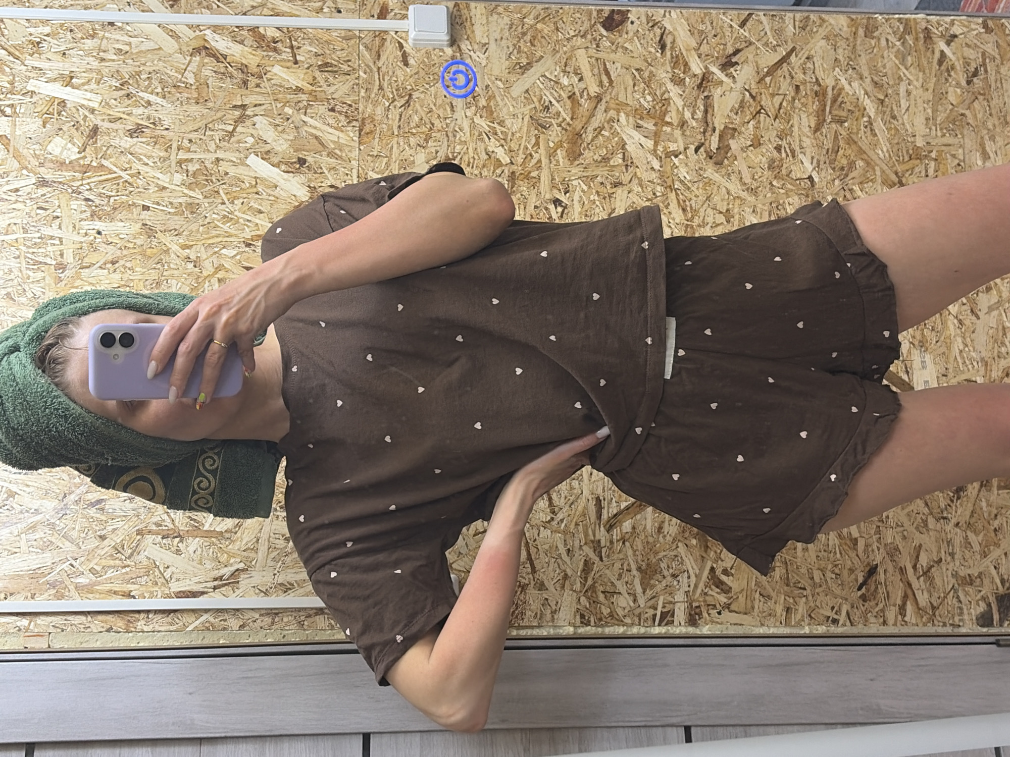 A woman stands in what appears to be a bathroom, taking a selfie in the mirror. She has a green towel wrapped around her head and is holding a light purple cell phone. She is wearing a brown pajama set with white heart patterns. The background behind her is a wall of particle board and a section of painted wall underneath. A light switch and decorative circular object are on the wall. Based on the presence of particle board and decorative elements, and the casual setting, this image is most likely from an interior location, but without other details, the exact location in Chaikovsky, Russia cannot be determined.