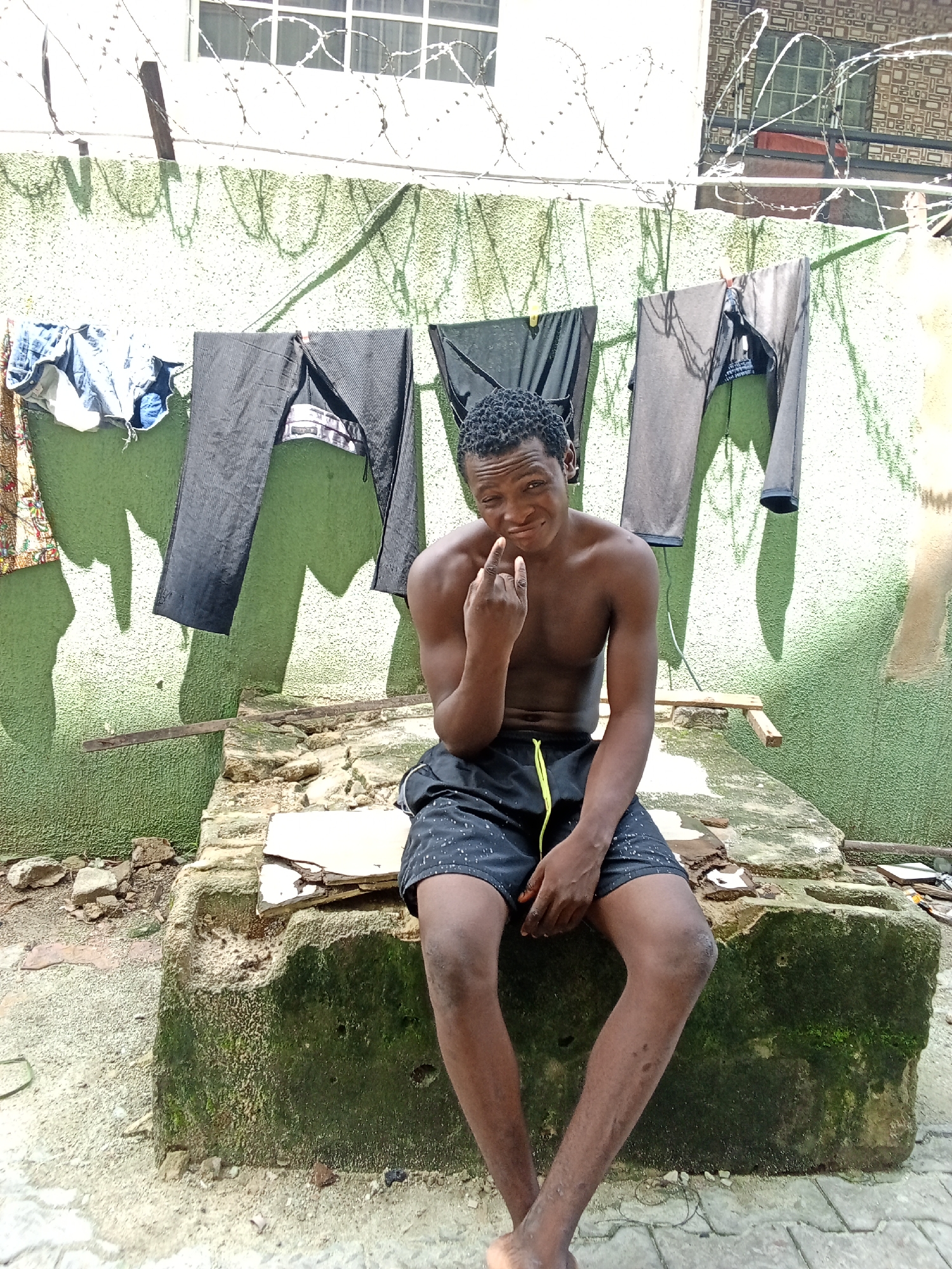 An outdoor scene in what appears to be a residential area of Ado, Nigeria, features a young Black male seated on a weathered concrete block. The individual, bare-chested and wearing black shorts with a yellow drawstring, faces slightly left of center with a subtle smirk, his left index finger raised. His lower legs display multiple small dark marks. Behind him, against a light green, rough-textured wall, several items of clothing (blue denim jeans, dark trousers) hang on a clothesline, casting distinct shadows. Above the wall, a barbed wire fence is strung, separating the foreground from adjacent buildings, one with white windows and another with a patterned facade. The concrete block the man sits on is discolored with moss and shows broken white tile fragments. The ground is unpaved earth with scattered debris and some grey pavers visible in the lower right. Bright daylight illuminates the scene.