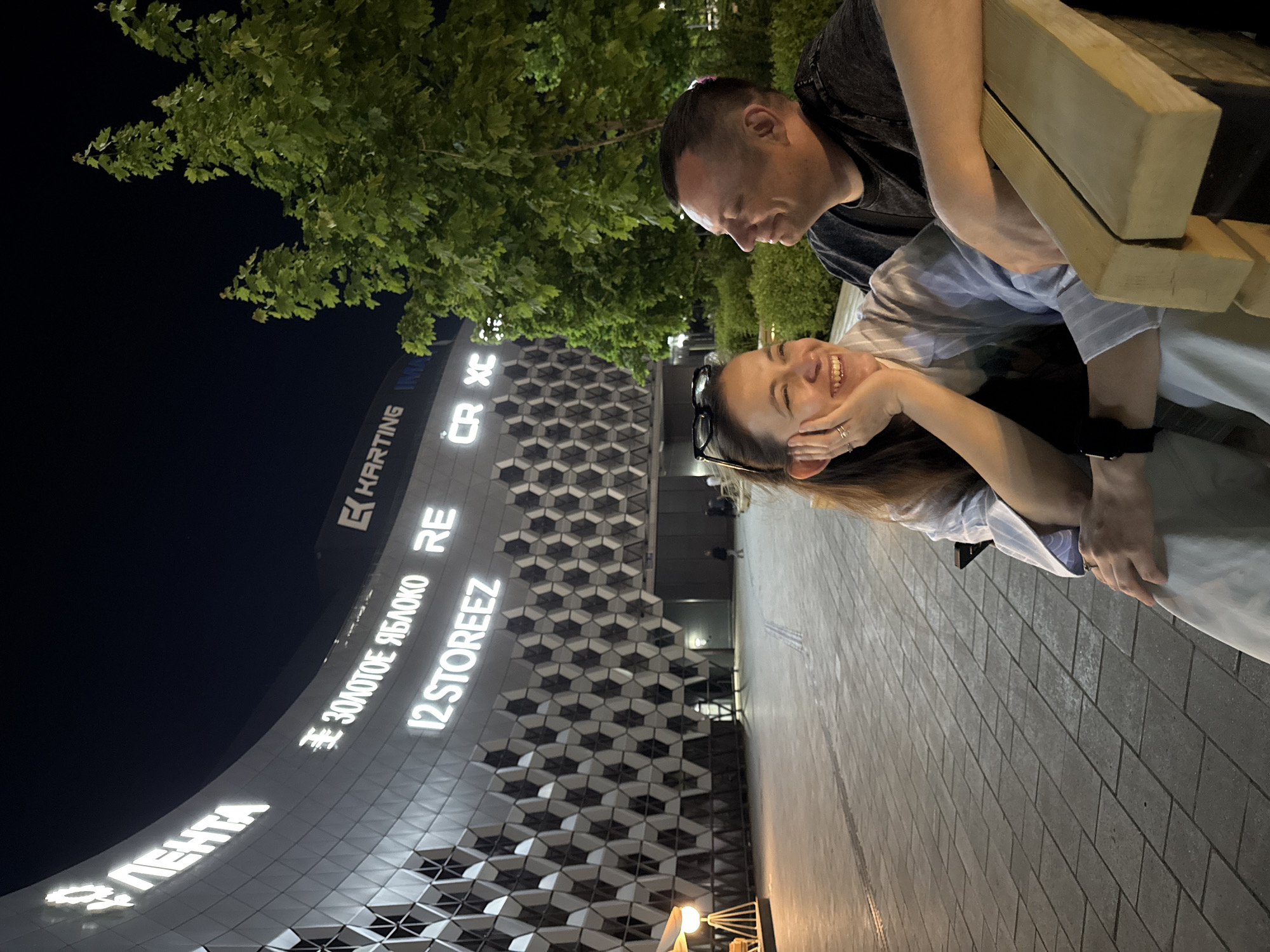 A medium shot captures a couple outdoors at night, with a large, illuminated building in the background. The building has a geometric design and several visible signs in Russian, including "ЛЕНТА" (Lenta), "#ЗОЛОТОЕ ЯБЛОКО" (#GoldenApple), "I2 STOREEZ," "RE," and "KARTING." A tree with dense foliage is visible above the couple.
The woman, positioned in the foreground, leans forward, her hand resting on her face, and smiles. Sunglasses are perched on her head. She wears a light-colored shirt with what appears to be a dark vest or layer underneath. A black watch is visible on her wrist. The man is slightly behind and to the right of the woman, looking down at her. His arm rests on a wooden structure, possibly a bench. He wears a dark shirt.
The setting appears to be a paved area in front of the building. Given the signs in Russian, the location is likely in Russia. Specifically, the presence of the "Golden Apple" store suggests this could be in Kazan, Russia. The image is taken at night, with the building's lights illuminating the scene.