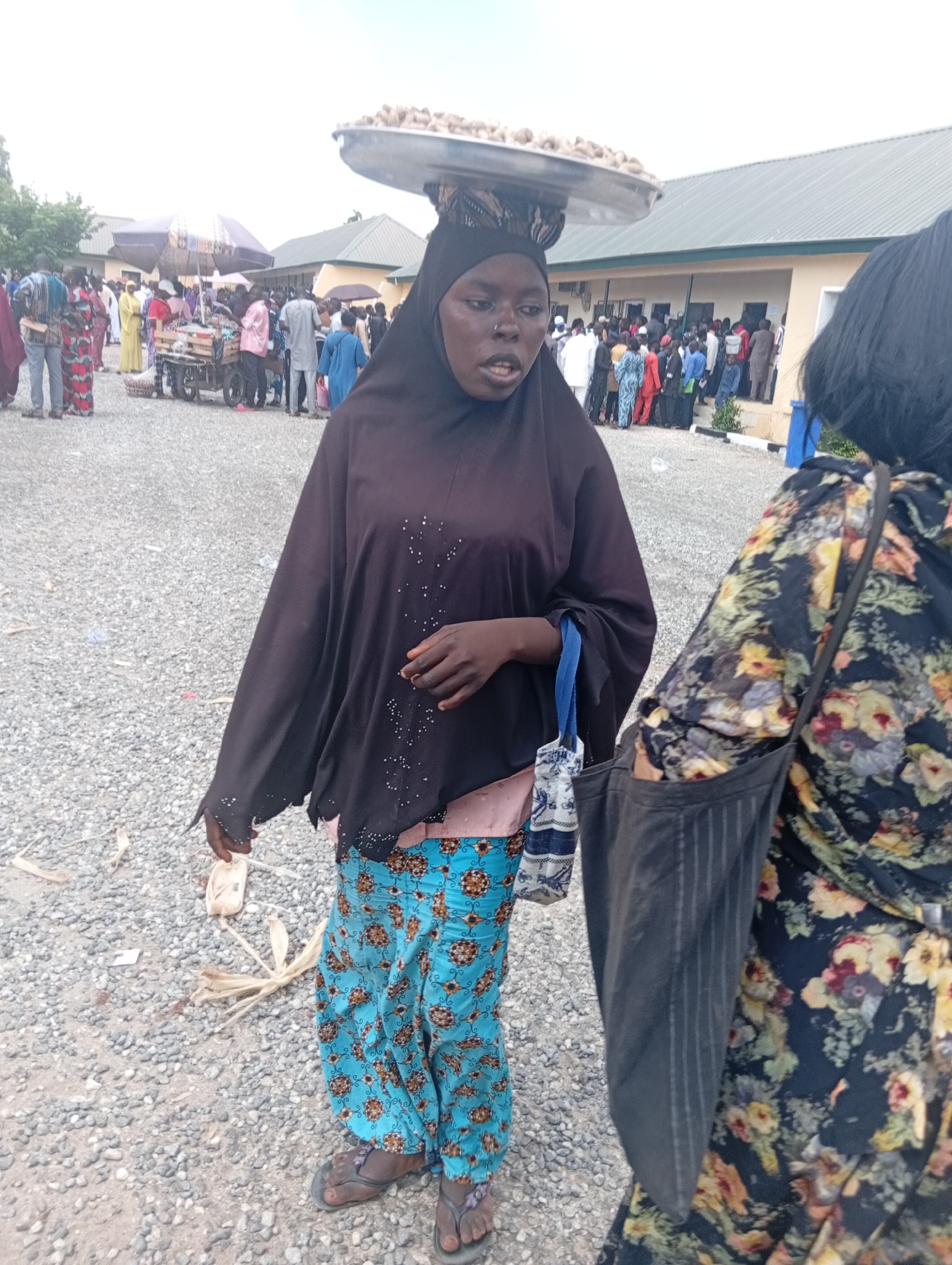 The photograph captures an outdoor scene, likely in Jalingo, Nigeria, where a woman is the primary subject. She is wearing a black hijab and a brightly patterned blue and floral print skirt. A tray filled with food, possibly nuts, is balanced on her head. She holds a small, white object in her right hand and a blue and white patterned bag hangs from her left arm. Behind her, a crowd of people are visible, some gathered around a vendor with a cart. The ground is covered with small gray stones. Another woman, partially visible on the right side of the frame, wears a floral print dress and carries a black bag over her shoulder. The background shows a building with a green roof. The lighting suggests it is daytime, with an overcast sky.