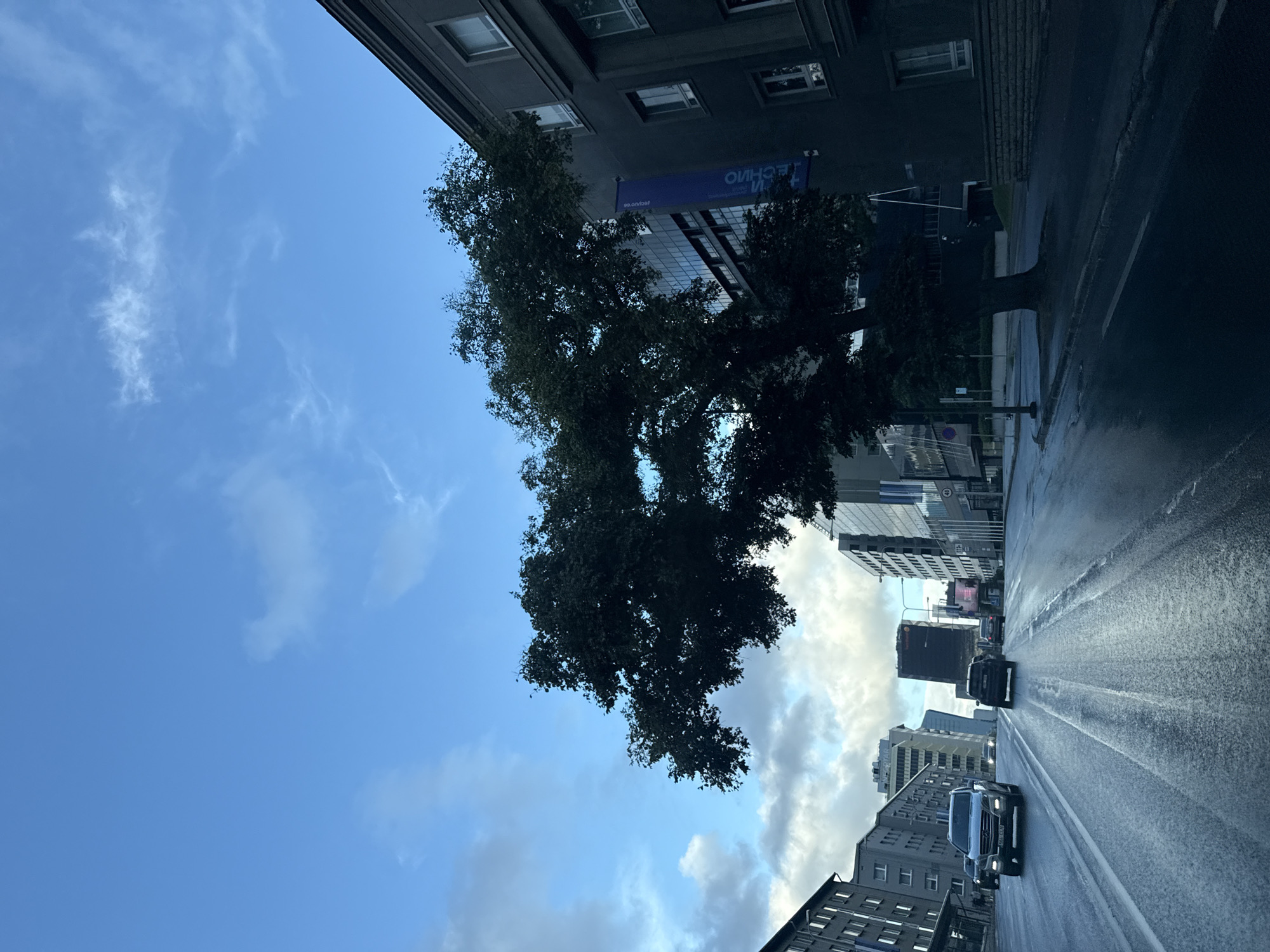 This is a street-level, first-person perspective shot taken from a vehicle traveling on a wet, multi-lane road in Tallinn, Estonia. The sky is a bright blue with scattered white clouds, indicating it is daytime. The road surface shows reflections of the sky and surrounding buildings, suggesting recent rain. Several vehicles are present on the road, with the closest being a silver SUV in the left lane and a dark SUV in the right lane, both moving away from the camera. In the distance, more cars and modern buildings of various heights and architectural styles line the street, some with glass facades.
Dominating the mid-ground on the right side of the frame is a large, mature tree with dark green foliage. Adjacent to the tree, a dark grey building with a facade of windows is visible. A blue banner with white text, appearing to be in Cyrillic or a similar script, hangs vertically from this building. Further down the street, a cluster of taller, contemporary buildings can be seen, including one with a distinctive angular shape. The overall impression is of a modern urban environment, likely a business or administrative district, under a clear but overcast sky.