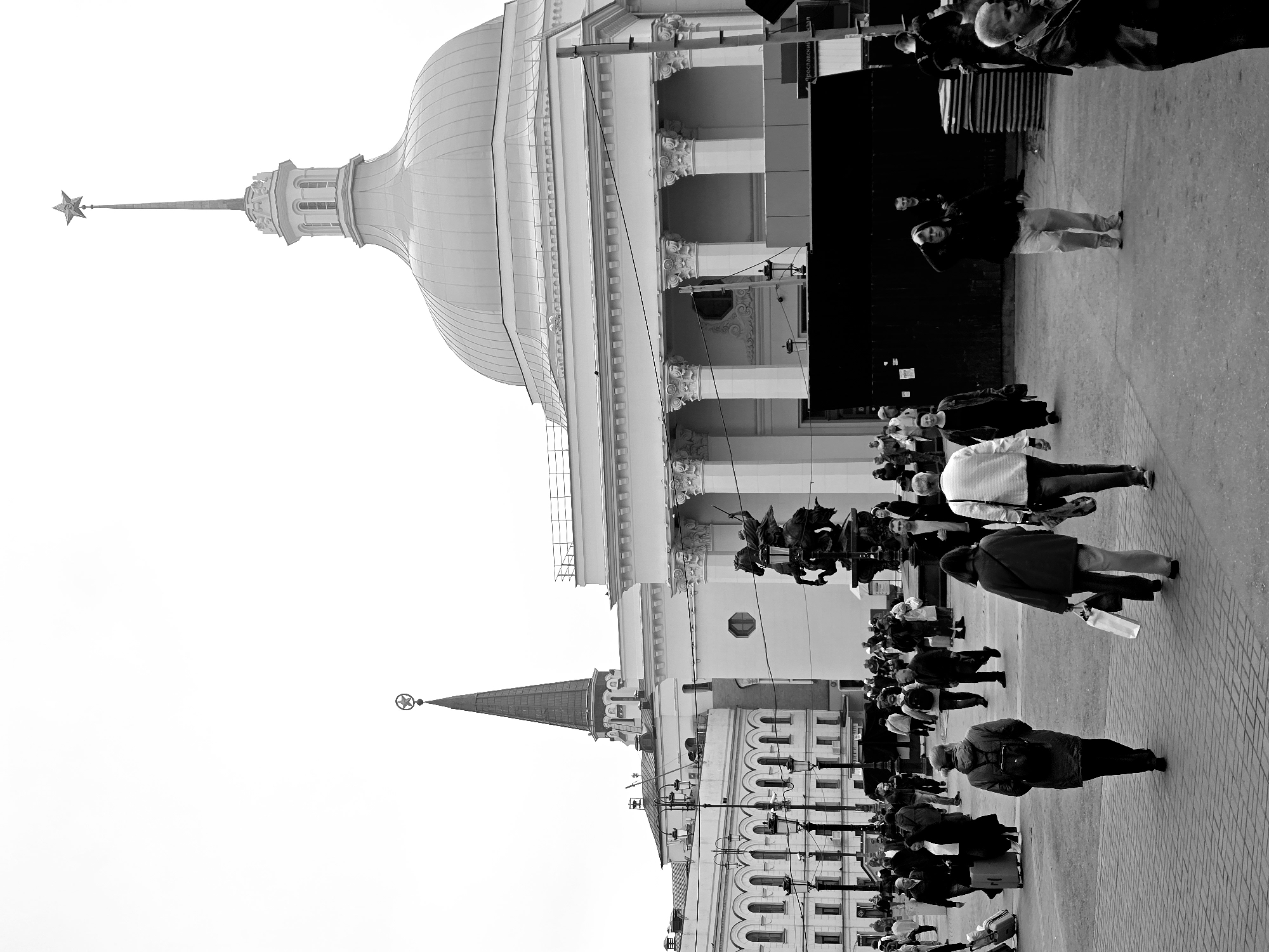 The image shows an urban scene in Moscow, Russia, featuring the architecture of the Red Square area. In the foreground, numerous pedestrians are walking on a paved area. In the background, there is a large building with a dome and a spire topped with a star. Another building with a tower and a spire topped with a star is visible on the left. The scene is in black and white.