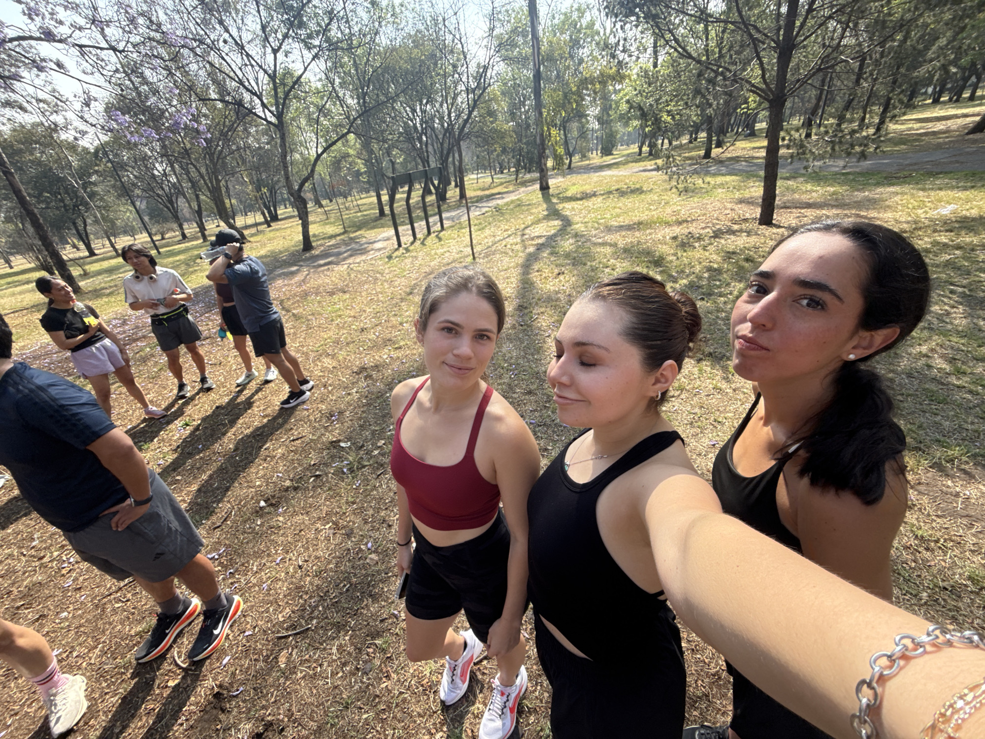 The image shows three women taking a selfie in a park in Mexico City. The location is likely Chapultepec Park, indicated by the large trees, dry grassy ground cover, and other individuals engaged in recreational activities in the background. The women are all wearing athletic attire, suggesting they may have been exercising. The woman on the left is wearing a red sports bra and black shorts, the woman in the middle is wearing a black tank top and shorts, and the woman on the right is wearing a black tank top. Several other people in athletic wear are in the background. The ground is covered in dry grass and some fallen leaves. The day appears to be sunny, casting long shadows across the grass.