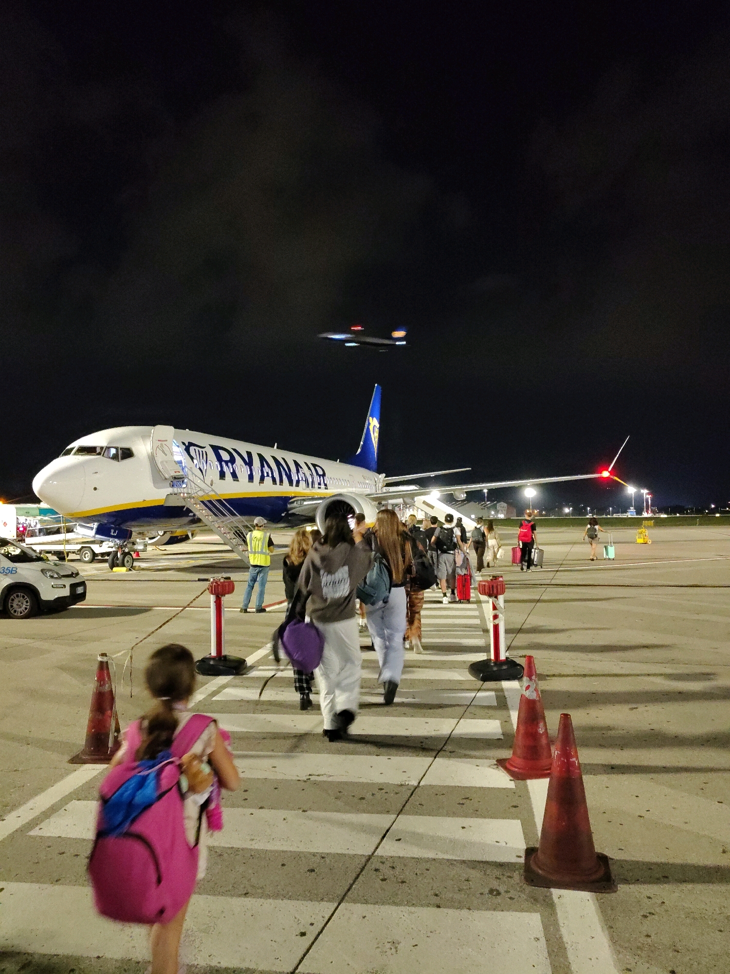 The image depicts a nighttime scene on an airport tarmac, with a large Ryanair airplane parked and boarding in progress. Several passengers are walking in a line towards the plane, with luggage in tow. A young girl in the foreground is walking away from the camera, wearing a bright pink backpack. In the background, another airplane is visible in the sky, possibly taking off or landing. The tarmac is illuminated by artificial lights, casting long shadows. The overall atmosphere is one of travel and movement, with the dark sky providing a stark contrast to the illuminated aircraft and people. The location is identified as Unknown, Unknown.
