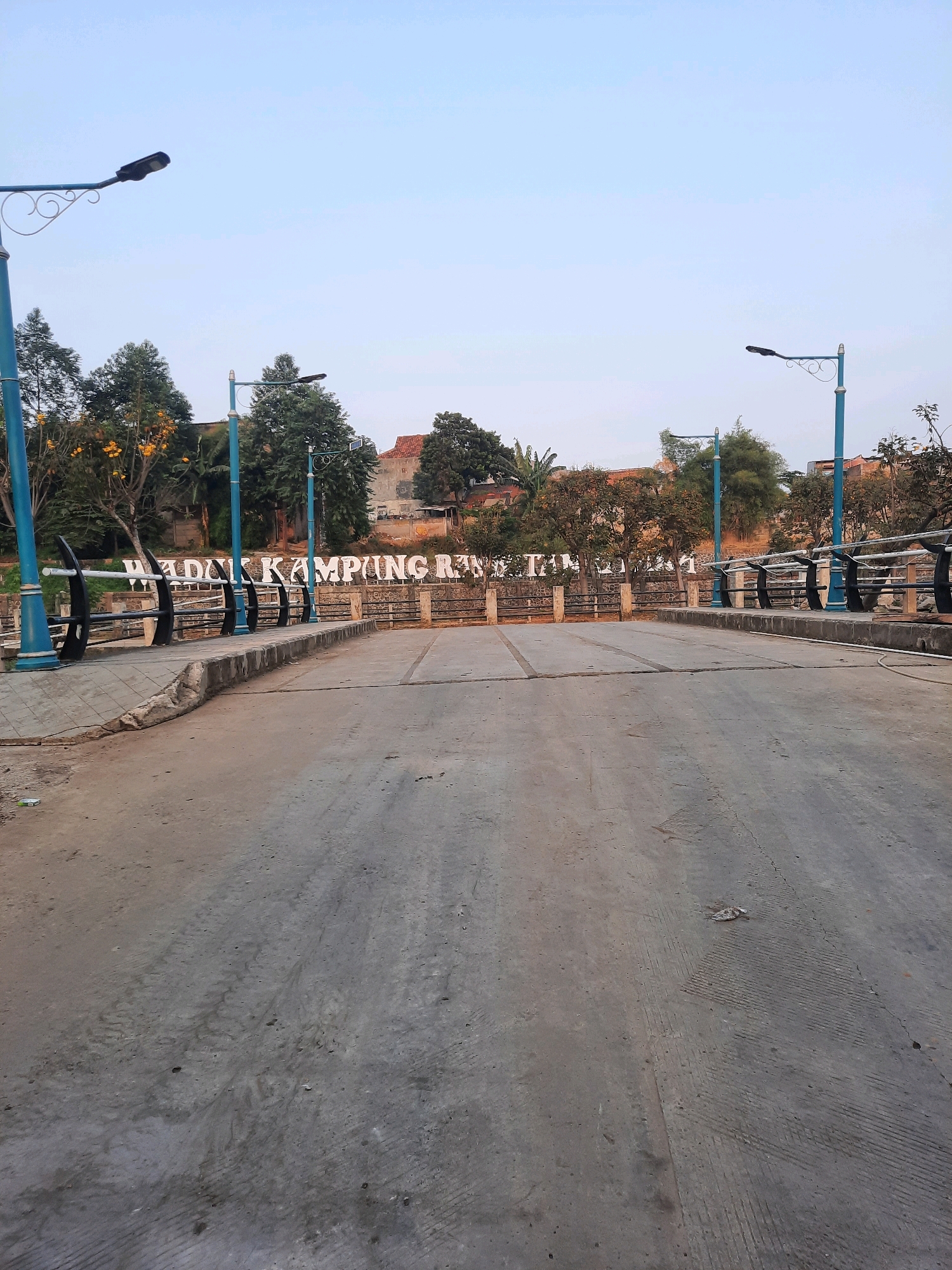 The image shows a straight, empty concrete road leading across a bridge. On either side of the bridge are decorative blue lampposts and black and silver railings. In the background, large white letters spell out "WADUK KAMPUNG RAKYAT". Behind the letters is a stone wall and beyond that, trees and buildings are visible. The sky is clear and pale blue, suggesting it is either early morning or late afternoon. The road appears to be recently paved or cleaned, with visible tire tracks and some small debris scattered on the surface. There are no people or vehicles in the scene. The location is identified as Waduk Kampung Rakyat in East Jakarta, Indonesia.