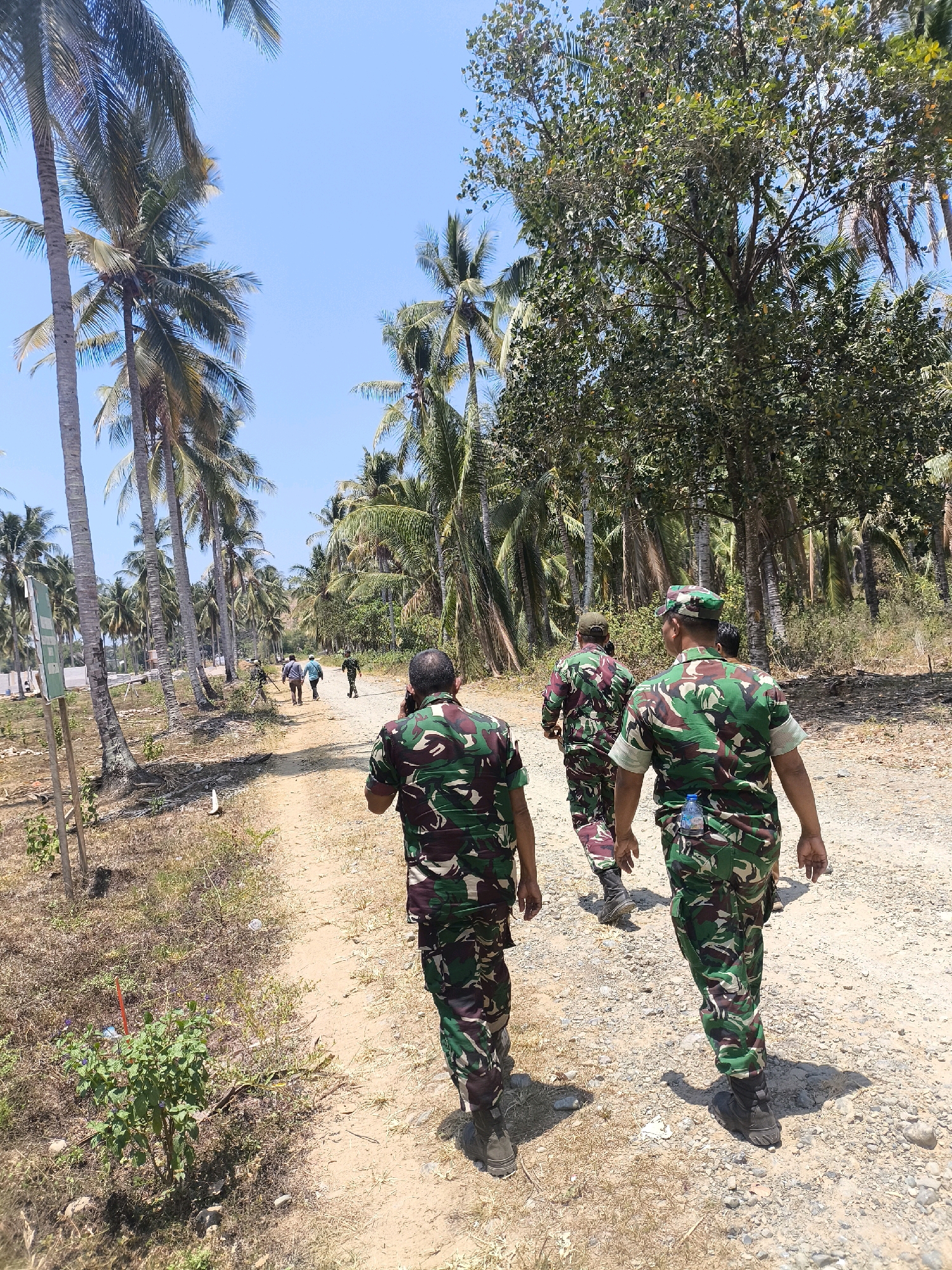 The image shows several individuals in military camouflage uniforms walking along a dirt path lined with palm trees and other vegetation. The sky is clear and blue, indicating a sunny day. In the foreground, a person in a camouflage uniform is facing away from the camera, holding a mobile phone to their ear. Behind them, three other individuals in similar uniforms are walking ahead on the path. Further down the path, a few more figures can be seen, along with what appears to be a white signpost on the left side of the image. The environment suggests a tropical or subtropical location. The presence of palm trees and the style of vegetation are consistent with a coastal area in Indonesia.