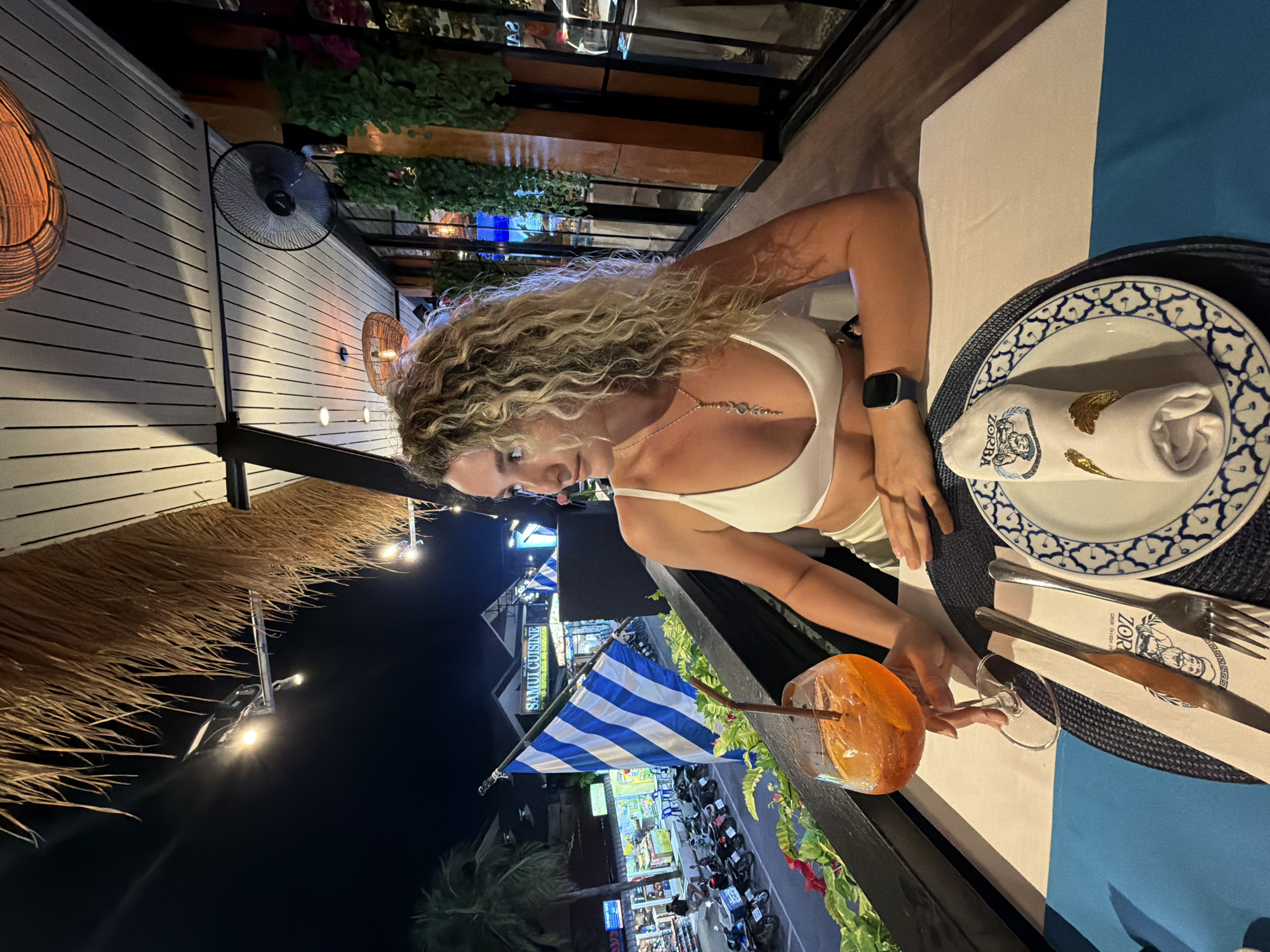 A woman with curly blonde hair sits at a table in a restaurant at night in Baan Chaweng, Thailand. She is wearing a white bikini top and a pendant necklace. In front of her is a glass of orange liquid with ice and a straw. The table is set with a plate, napkin, fork, and knife. In the background, a street scene with shops and parked motorcycles is visible, along with a flag that reads "SAMUI CUISINE". The restaurant has a thatched roof and a ceiling fan.