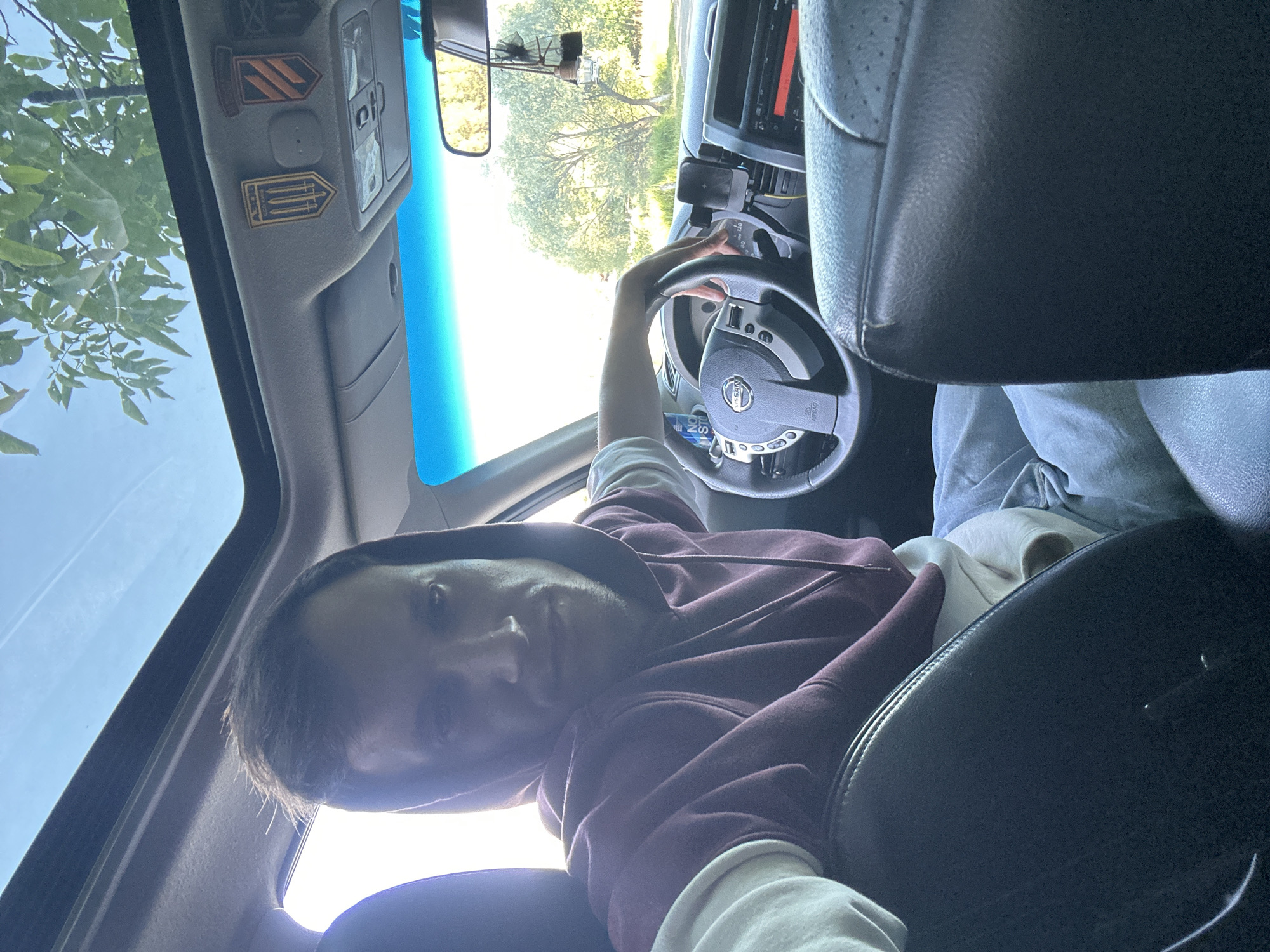 A man wearing a maroon hoodie with a white undershirt and light blue jeans is sitting in the driver's seat of a car. The car has a sunroof open, revealing a bright sky and green trees. On the ceiling of the car, near the sunroof, are two patches, one with a yellow shield and three arrows, and another with orange and black diagonal stripes. The man has his left hand on the steering wheel, and his right arm is resting on the dashboard. The steering wheel is dark gray with silver accents, and it has "SRS AIRBAG" written on it. A black phone mount is attached to the dashboard. The interior of the car appears to be leather. The background outside the car shows trees and a bright, sunny day. The image is a selfie taken from inside the car, looking towards the driver. There are no obvious city landmarks visible to confirm the location in Makiv, Ukraine.