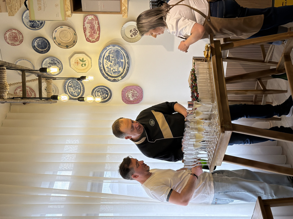 The image portrays an indoor scene within a modern establishment in Felgueiras, Portugal, set up for a social gathering or tasting event. Three individuals are positioned around a rectangular wooden table.
To the left, a male subject in a white short-sleeved t-shirt and light blue jeans is opening a dark green bottle over the table. In the center, a male subject, identified by a black chef's jacket with a beige diagonal stripe and a "Chocolate" logo, stands looking down at the table's contents. To the right, a female subject with shoulder-length grey-blonde hair, wearing a white short-sleeved blouse, dark jeans, and a brown apron, holds a small metallic plate with food.
The table is laden with multiple clear glass champagne flutes, most filled with a pale yellow liquid. Adjacent to the glasses is a rectangular white dessert, likely a cake, topped with an arrangement of red berries, blueberries, and green leaves. A small dish with a pastry or snack is visible in front of the female subject. A green bottle rests on the floor beneath the table.
The background features a large window on the left, covered by floor-length translucent white vertical blinds. The white wall is decorated with an array of antique-style decorative plates of various sizes and patterns, predominantly in blue and white, red, and pink. Above the plates, a chandelier-like fixture constructed from a wooden ladder-like frame suspends multiple exposed filament light bulbs. On the right, a light-colored wooden shelving unit displays packaged goods, with visible text including "SÃO LUIZ Winemaker" and "Quinta Nova."