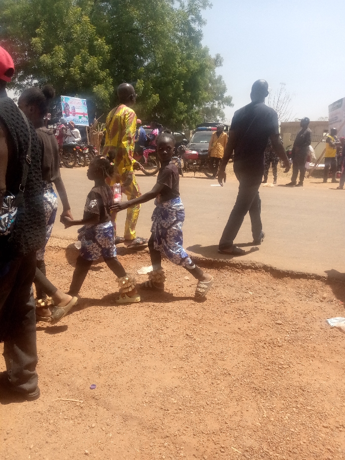This image depicts a street scene in Kagoma, Nigeria, under bright daylight. In the immediate foreground, two children are walking right-to-left across a reddish-brown dirt and gravel area adjacent to an asphalt road. The child on the right, appearing slightly older, wears a black short-sleeved shirt, blue and white patterned wraparound fabric around the waist, light-colored sandals, and decorative ankle adornments resembling white shells. This child is looking directly towards the camera. The second, smaller child to their left wears a similar black shirt, blue and white patterned wraparound fabric, light-colored sandals, and identical ankle adornments. This child is holding the hand of the first child. Partially visible to the far left is a female figure in a black top and similar patterned skirt, holding the hand of the smaller child.
Behind the children, a man in a yellow and black patterned long-sleeved shirt and trousers walks away from the camera. To the right of the children, another man in a black short-sleeved shirt and dark trousers walks away from the camera, with his back partially to the viewer. The background features several individuals, some on motorcycles, and other vehicles, including a dark-colored vehicle with what appears to be a light bar on its roof. A blue and white sign is visible amongst green foliage on the left side of the background. The sky is clear and bright. The ground transitions from uneven dirt and gravel to a darker asphalt road. Scattered litter, including a purple bottle cap and white paper, is visible on the dirt.