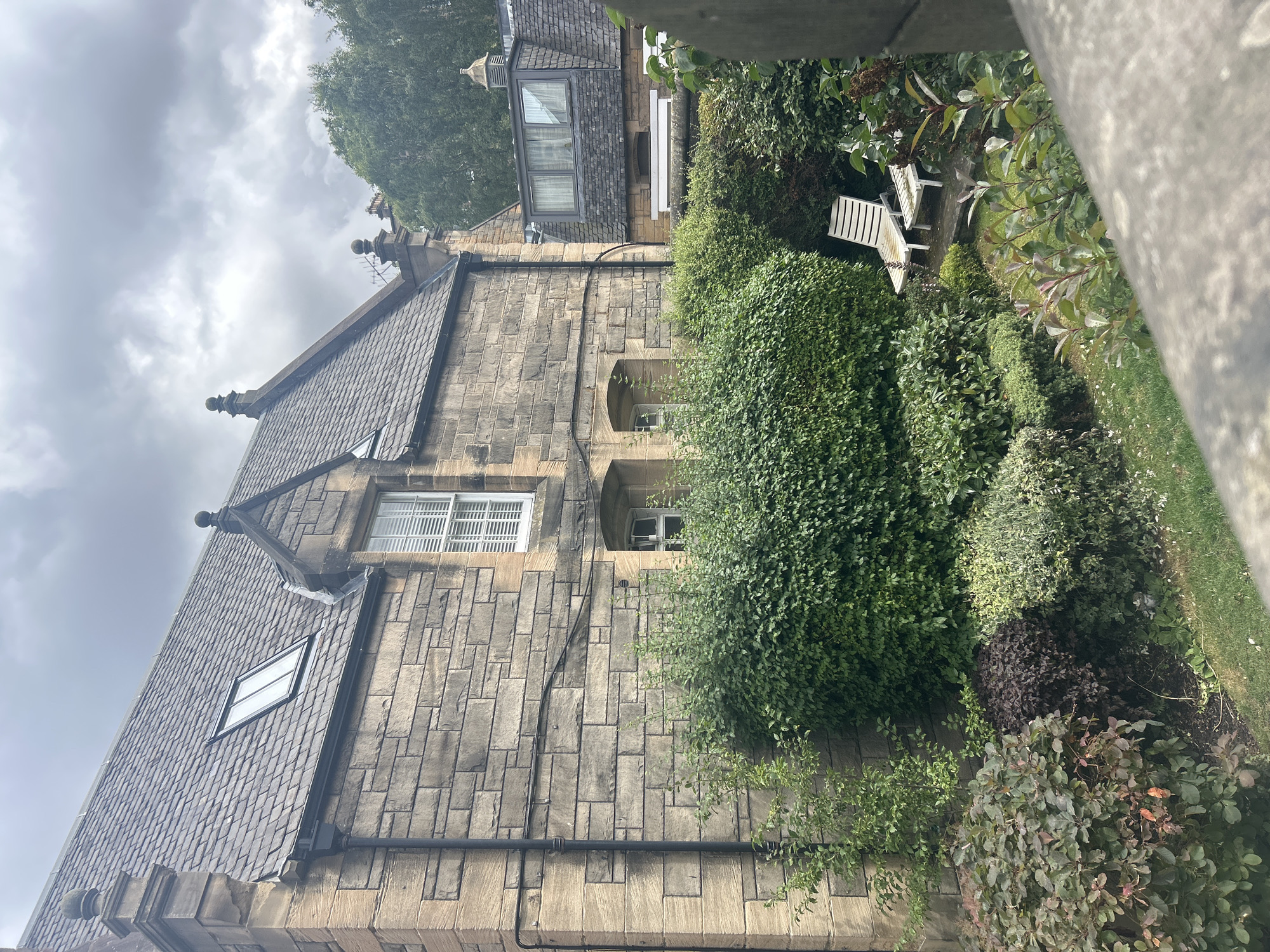 The image depicts a view of a stone building with a slate roof, characteristic of architecture found in Edinburgh, Scotland. The building features a dormer window and multiple smaller windows. In the foreground, a lush garden with various shrubs and manicured bushes is visible. Two white lounge chairs are placed on a small patio within the garden. The sky is overcast with clouds. The overall scene suggests a residential property in a well-established neighborhood within the city.