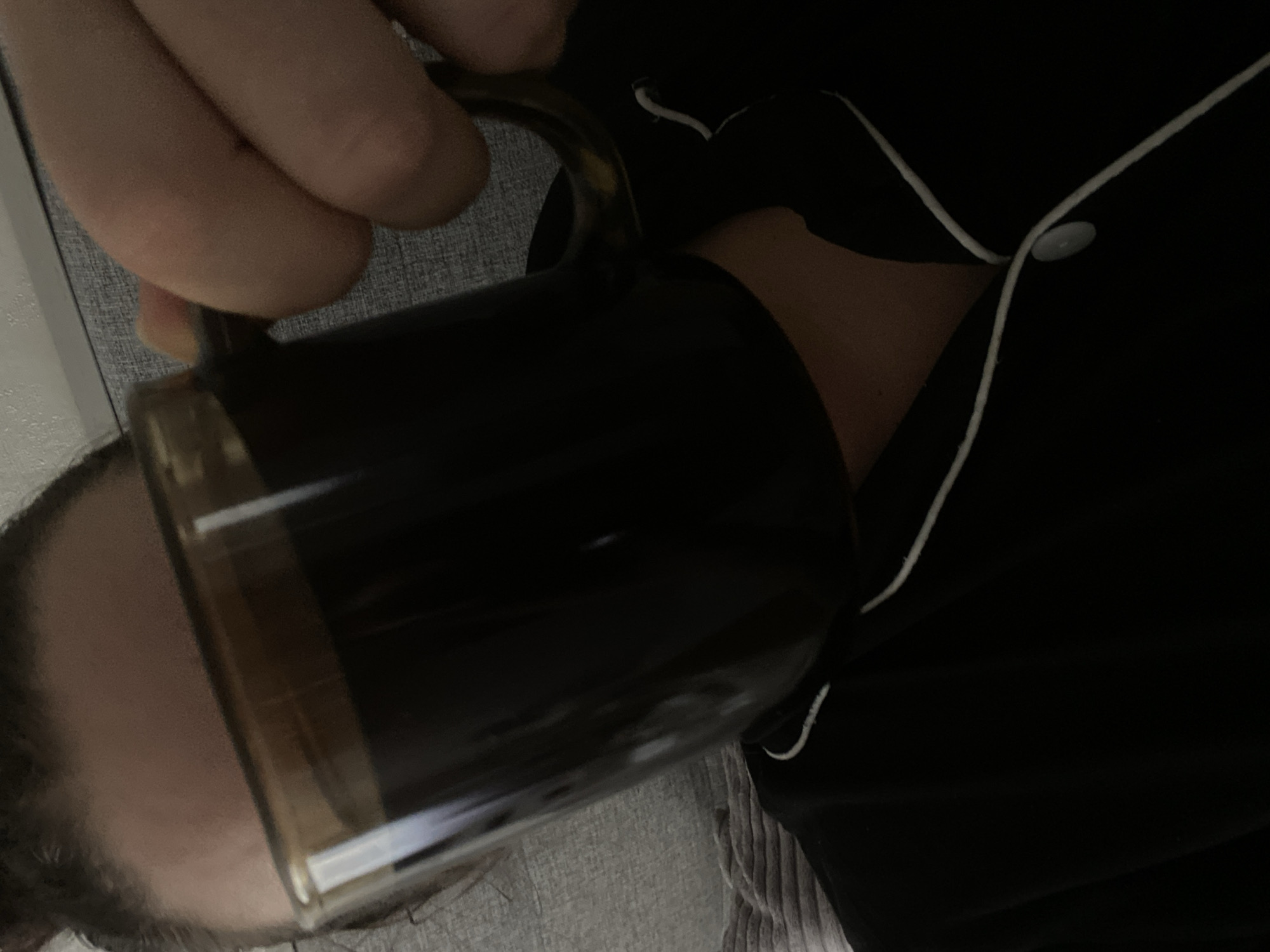 A person is holding a dark-colored liquid, likely coffee or tea, in a mug. The person is wearing dark pajamas with white piping along the collar and front opening. The scene appears to be indoors, possibly in a home or hotel room. The background shows a gray, textured surface, possibly a sofa or bed. There's no information that indicates the location is in the city of Sochi, Russia.