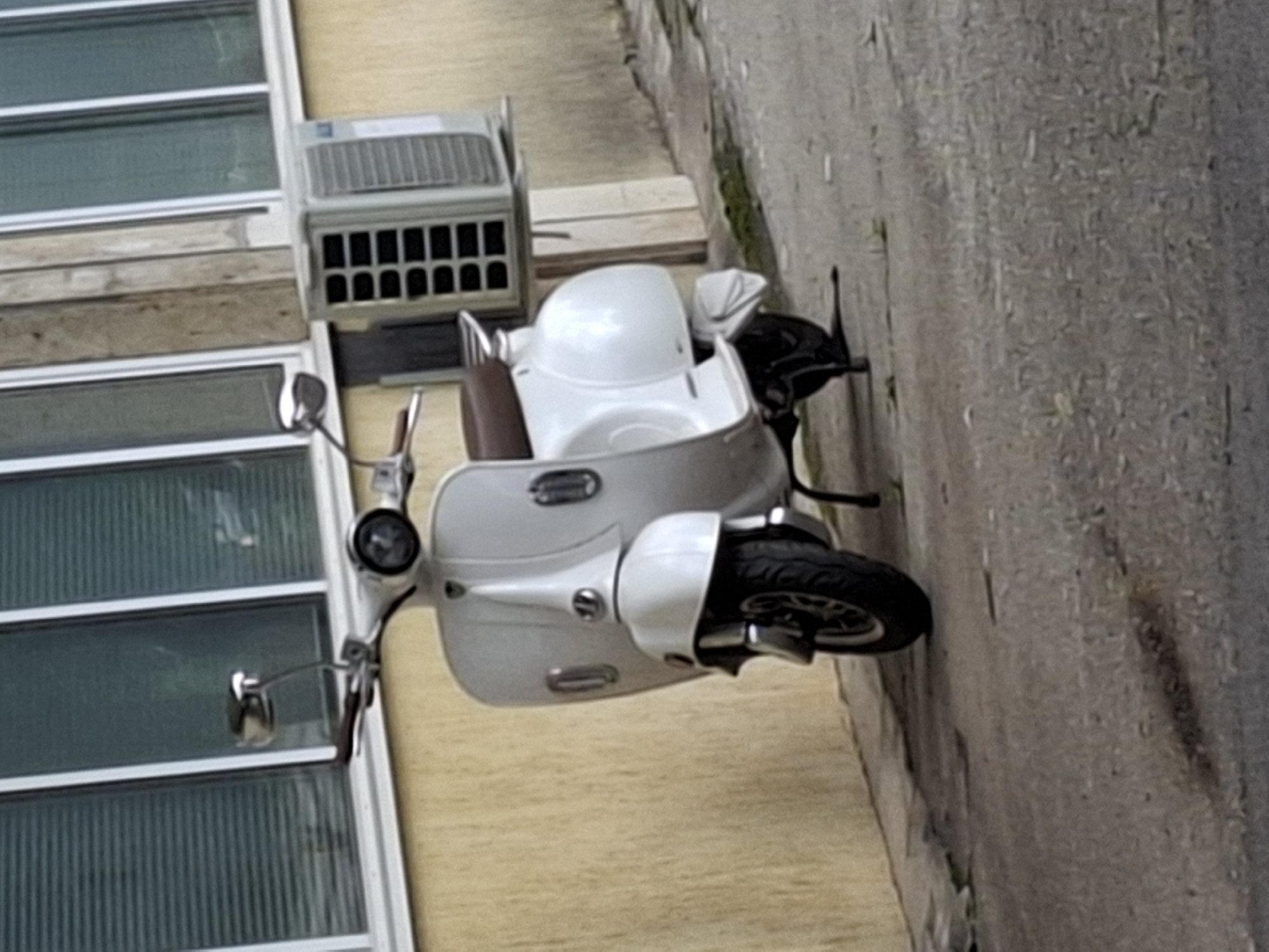 A white, vintage-style scooter is parked on a grey concrete or asphalt pavement next to a light yellow building. The scooter features a brown seat, chrome handlebars with rearview mirrors, and a single front headlight. An external air conditioning unit, primarily white and grey with a distinctive rectangular gridded vent, is positioned on a flat, light-colored support structure over the scooter's rear wheel, appearing to be affixed to its frame. The building behind the scooter has a textured, light yellow or beige facade and multiple windows with white frames, behind which horizontal blinds are visible. A vertical light brown pillar or beam is situated between two windows on the right side of the frame. The pavement shows some unevenness, minor cracks, and sparse patches of green vegetation or moss. Shadows are cast on the ground, indicating an overhead light source. No individuals are present in the scene, and no specific actions or interactions are depicted. The general location is Sofia, Bulgaria, though no specific landmarks or street indicators are visible to pinpoint an exact address within the city.