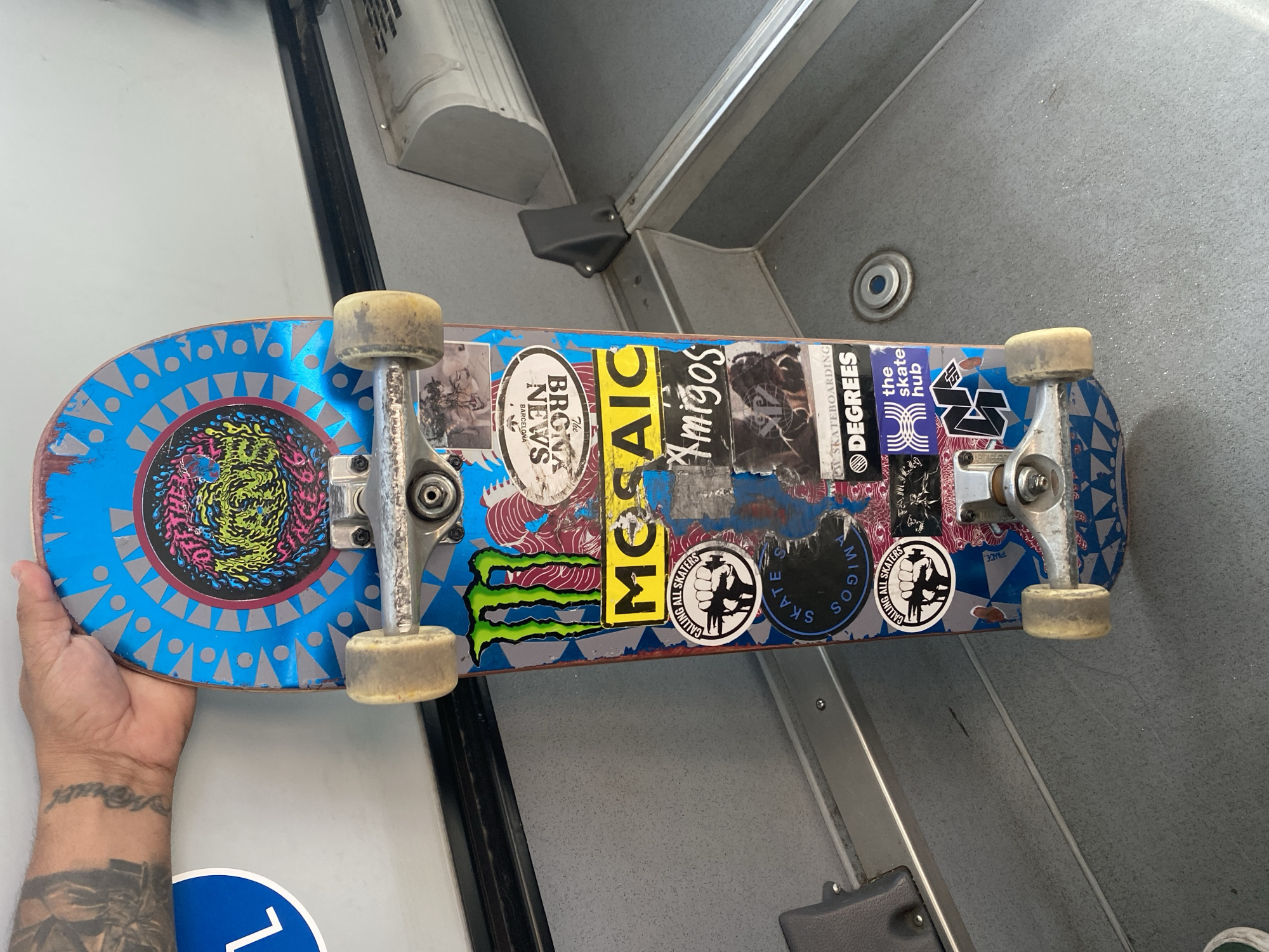A close-up image shows a heavily stickered skateboard being held by a person's left hand and forearm, revealing multiple tattoos including text on the wrist. The skateboard deck is vibrant blue with a detailed grey and silver geometric pattern, surrounding a central, colorful graphic that appears to be a psychedelic monster or abstract design in pink, yellow, green, and black. The skateboard features worn silver trucks and off-white wheels.
Numerous stickers adorn the skateboard's surface, some overlapping. Visible text and logos include "MOSAIC," "Amigos," "DEGREES SKATEBOARDING," "the skate hub," the "Monster Energy" logo, and two circular stickers reading "CALLING ALL SKATERS" with a graphic of a raised fist holding a skateboard. Notably, a "BRONA NEWS BARCELONA" sticker indicates the location context of Barcelona, Spain. A partially visible "VANS" sticker can also be seen.
The setting appears to be indoors, possibly within a public transport vehicle like a bus or train, given the grey, textured, and ribbed floor, metal railings, and utilitarian design elements in the background. A small blue and white circular sticker with the number "1" is partially visible on the floor near the person's arm. The time of day and weather are not discernible, and no specific activity beyond holding the skateboard is clearly depicted.