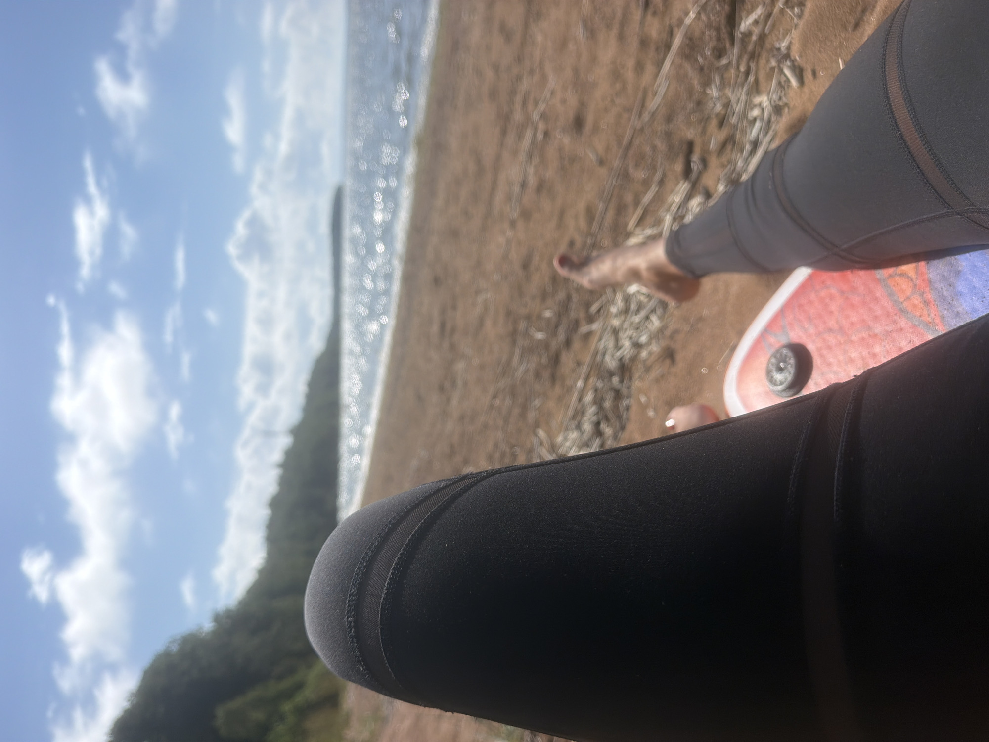 The image shows a close-up view of a person's legs, clad in black leggings, resting on a sandy beach. The person is lying down, with their feet visible towards the bottom right of the frame. A portion of a red and white object, possibly a board, is under their legs. The background depicts a calm body of water, a sandy shore, and a tree-covered hillside under a cloudy blue sky. The scene suggests a relaxing day at the beach. The location is the city of Vyazy, Russia.