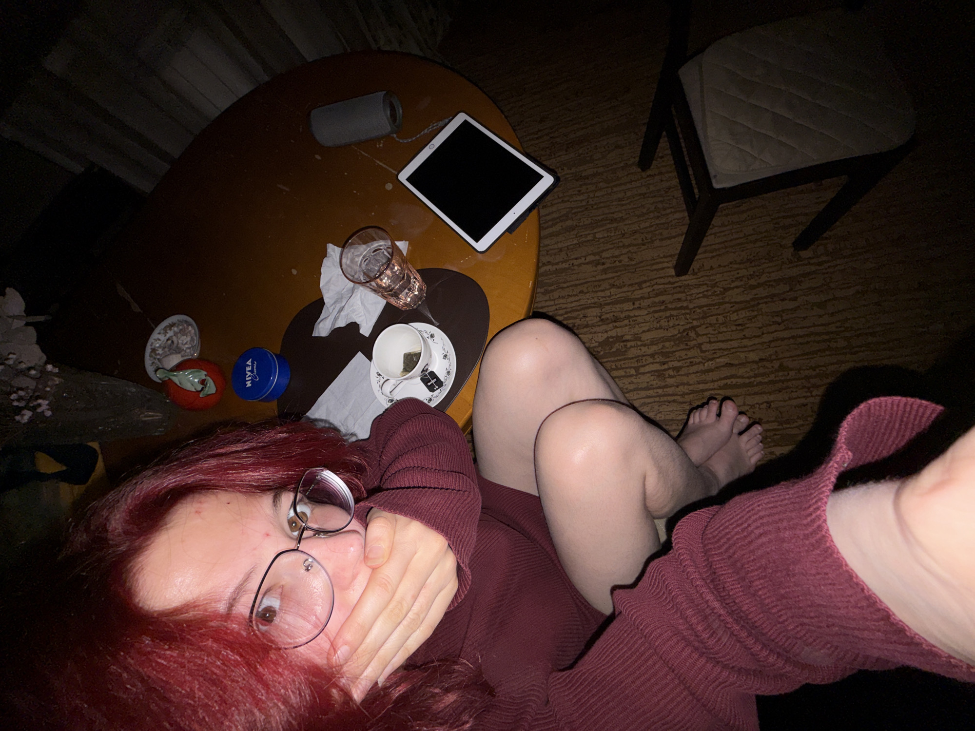 A person with bright red hair and glasses is sitting on the floor in front of a round wooden table. They are wearing a maroon sweater and have their hand covering their mouth. On the table are a cup of tea, a glass of water, a Nivea cream container, a tablet, and a portable speaker. A chair is lying on its side on the floor next to the table. The room appears to be dimly lit, possibly at night, and the flooring is a patterned linoleum. There are no specific indicators to place this scene in Chelyabinsk, Russia.