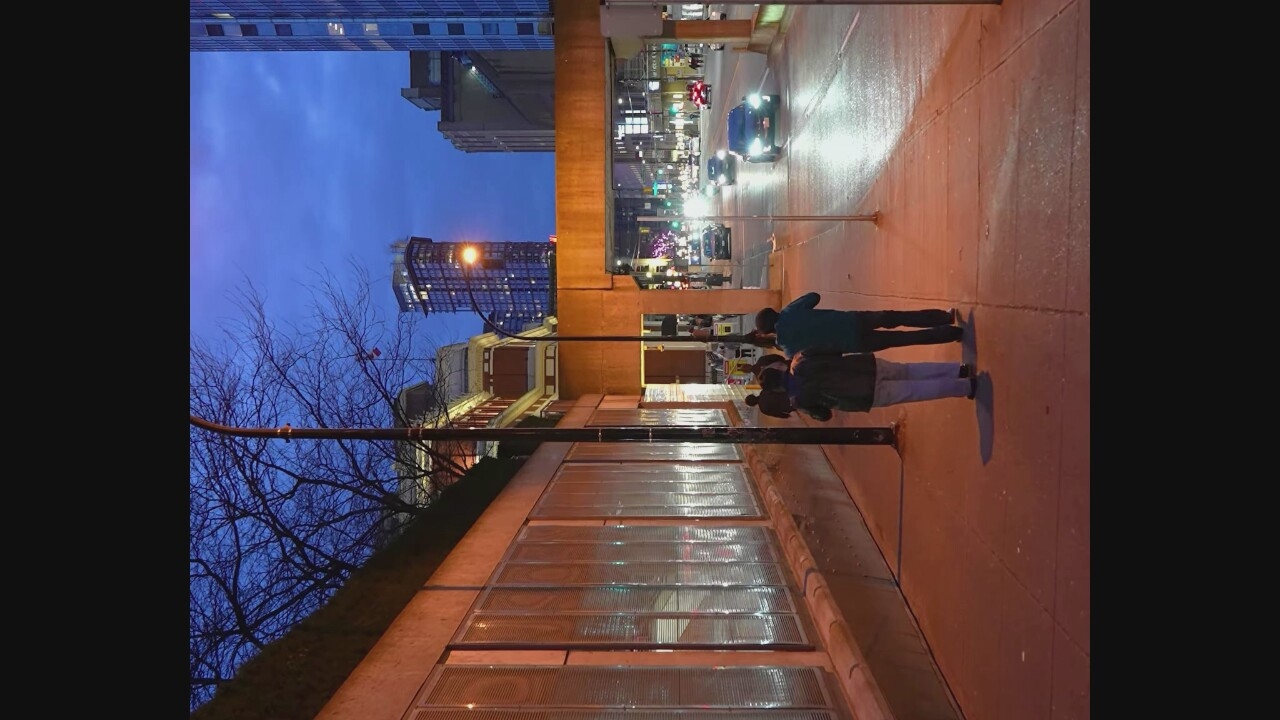 The image depicts a street scene at dusk in Vancouver, Canada, likely near a downtown area given the presence of tall buildings and illuminated city lights. In the foreground, two individuals are walking away from the camera on a sidewalk. The person on the left wears a dark jacket and light-colored pants, while the person on the right wears a dark jacket and dark pants. To the left of the sidewalk is a concrete wall with large, rectangular translucent panels that reflect light. A lamppost stands on the sidewalk between the wall and the street.
The street itself is wet, suggesting recent rain, and is illuminated by streetlights and the headlights of passing cars. Several vehicles are visible on the street, including a dark sedan in the right lane. In the background, a pedestrian overpass spans the street. Beyond the overpass, a variety of urban buildings are visible, including a prominent tall skyscraper with many illuminated windows. The sky is a deep blue, indicating the transition from day to night. The overall atmosphere is one of an urban environment at twilight, with a mix of artificial and natural light.