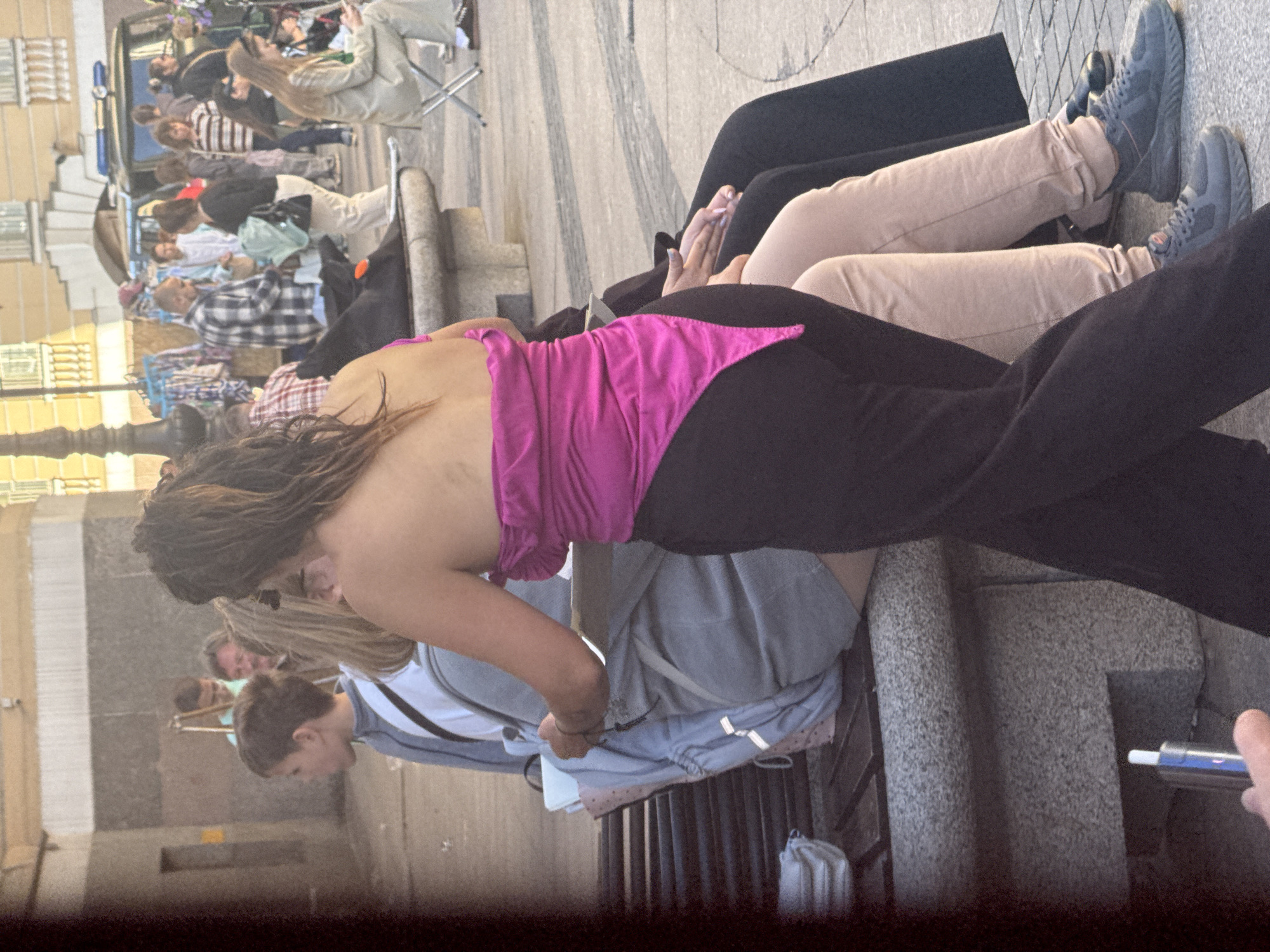 The image shows a street scene in Saint Petersburg, Russia, likely near Nevsky Prospect given the architecture. In the foreground, a person wearing a bright pink strapless top and black pants is standing. They are interacting with another person sitting on a stone bench, who is wearing a grey jacket over a light blue top and has a backpack. The standing person appears to be holding something and leaning towards the seated person. Behind them, other people are visible, some browsing at market stalls set up on the street. A white van with blue flashing lights is parked in the background, suggesting a public space. The lighting indicates it is daytime, possibly late afternoon, with soft shadows.