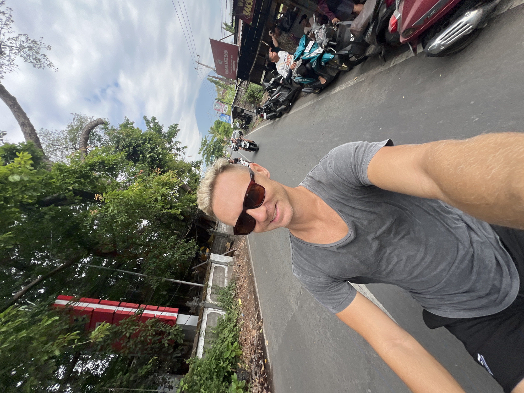 A man with blonde hair and sunglasses is riding a motorbike on a street in Abang, Indonesia. He is wearing a grey t-shirt and black shorts. In the background, there are trees, buildings, and other motorbikes. A volcano is visible in the distance. The sky is cloudy.