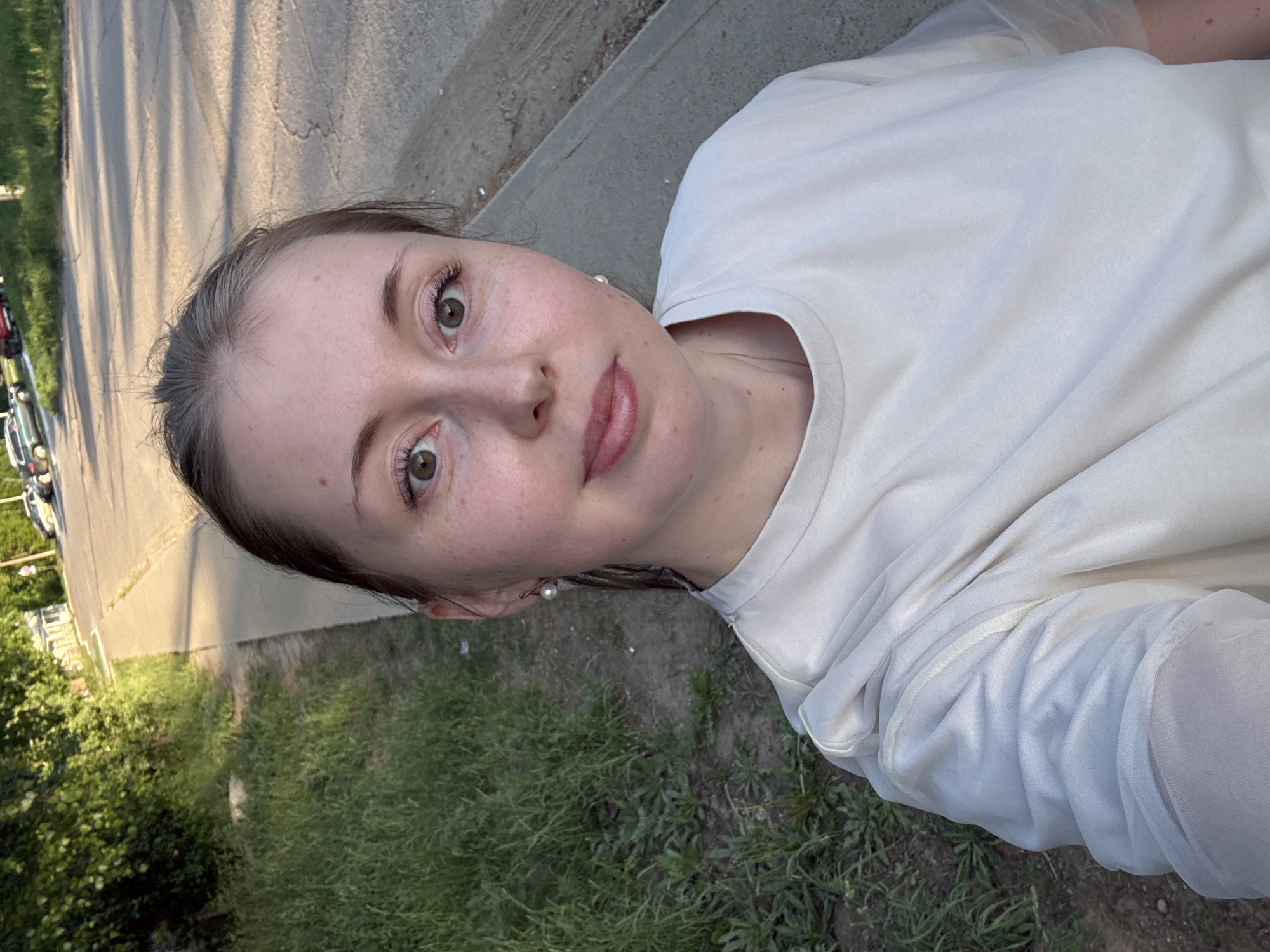 A woman with light skin, hazel eyes, and light brown hair pulled back, is taking a selfie outdoors. She's wearing a light-colored T-shirt and pearl earrings. The background includes a paved street, a concrete curb, green grass, and trees. Parked cars are visible in the distance on the left side of the image.