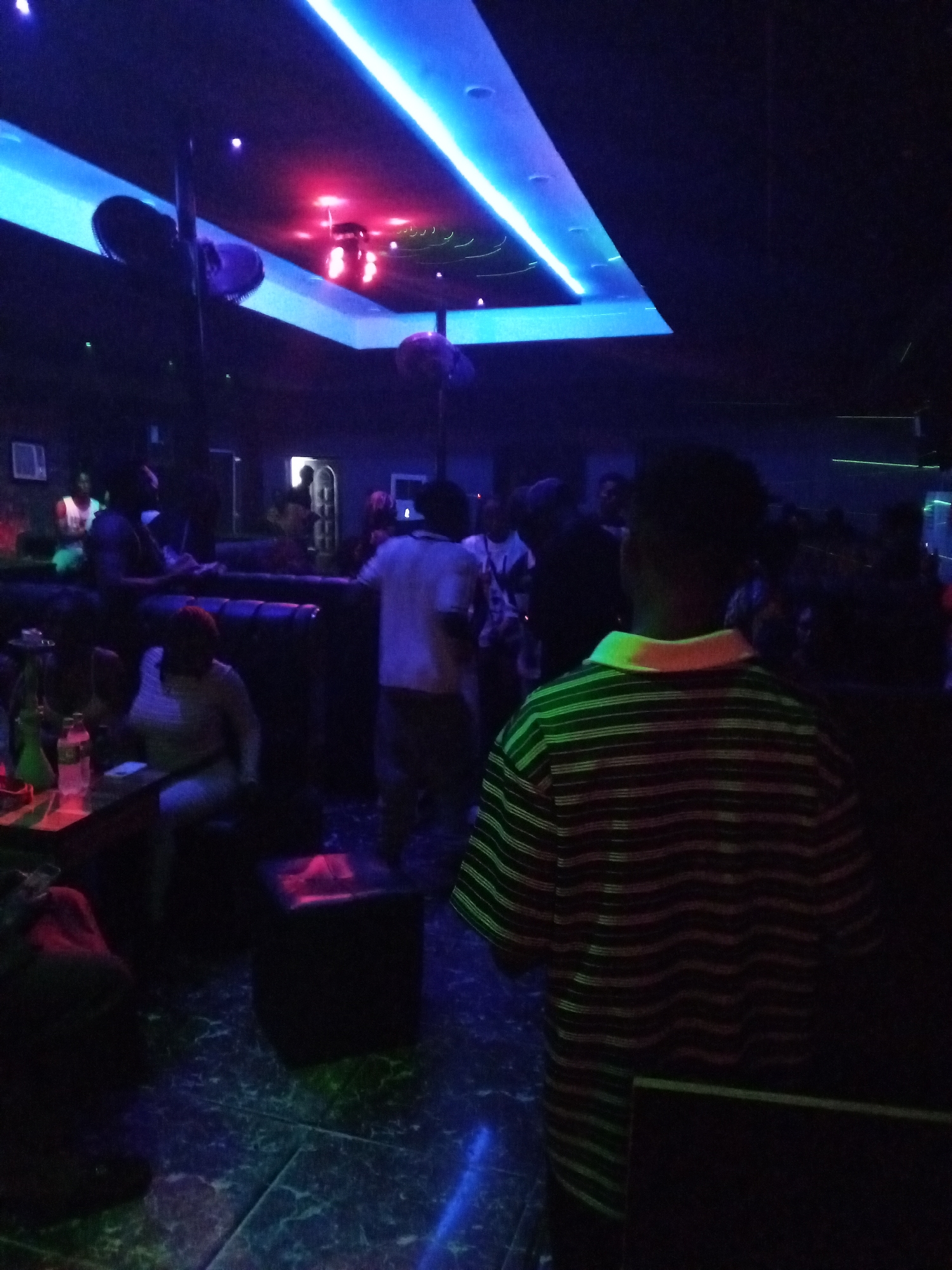 This media file depicts an indoor scene at night, likely within a club or lounge in Lokoja, Nigeria. The environment is dark, predominantly lit by neon light fixtures. A prominent feature of the ceiling is a long, bright blue LED strip light, running along an architectural recess. In another section, red lights illuminate a different part of the ceiling, possibly highlighting fans or decorative elements, which appear dark and indistinct.
Numerous individuals are present, most of whom are obscured by the low light. In the foreground, a person is seen from behind, wearing a striped polo shirt with a light-colored collar. Deeper into the room, several other people are visible. Some are seated on dark, upholstered benches or couches, forming seating areas that line the walls. One person is seated at a table where a hookah or shisha pipe and a plastic bottle are discernable. Other individuals are standing, appearing to socialize. The floor is tiled, reflecting some of the ambient blue and red lighting.
The activity taking place appears to be a social gathering or an evening out at an entertainment venue. The overall mood is relaxed, with people congregating and interacting. No specific texts are clearly visible in the image.