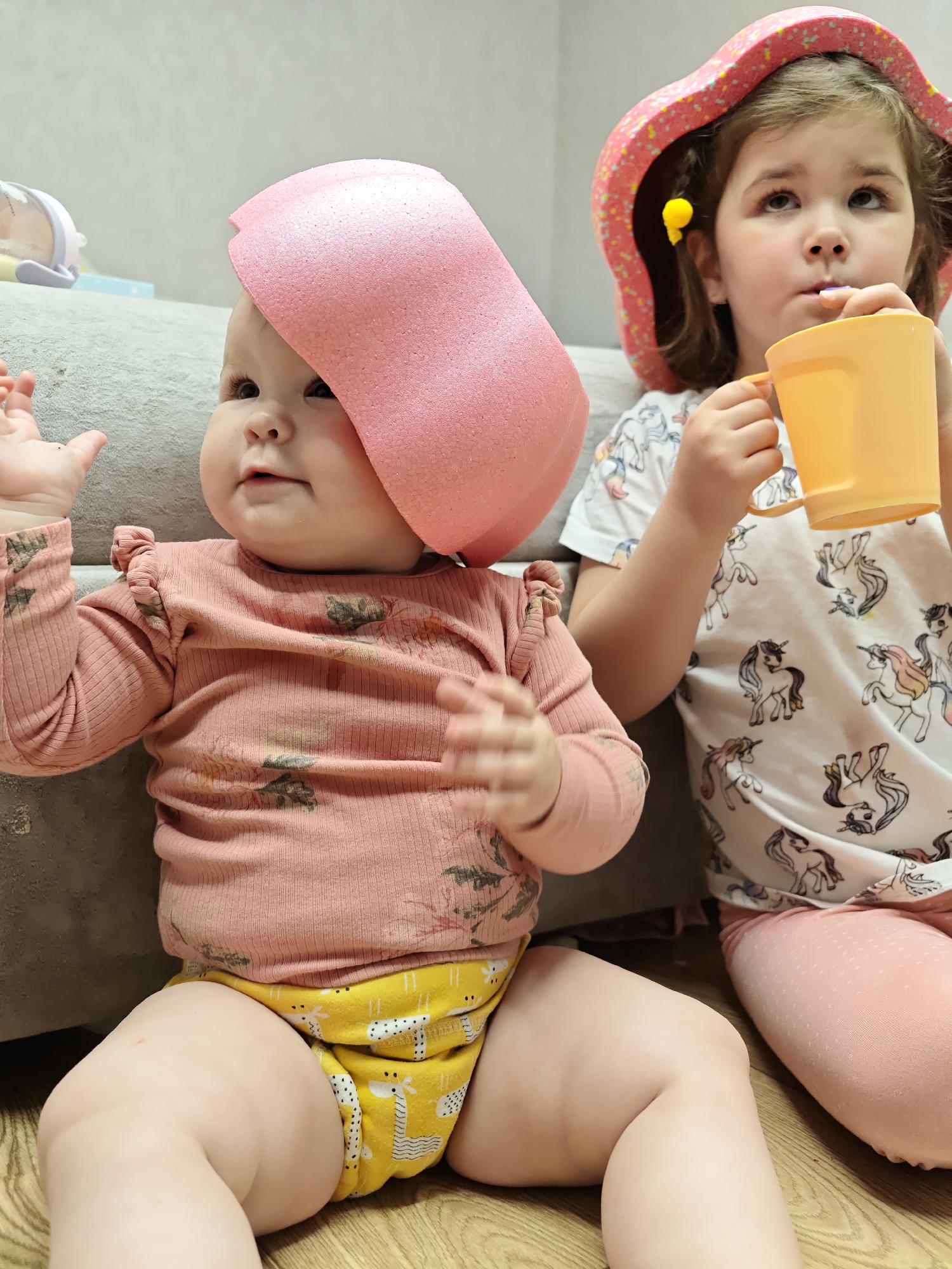 Two young children are seated on a wooden floor, seemingly indoors. One, a baby, sits with a pink foam object on its head, wearing a pink shirt with a floral pattern and a yellow diaper with a giraffe print. The baby's arms are raised with its hands open. Beside the baby, a slightly older child is holding a yellow mug, possibly drinking from a straw, and is wearing a pink sun hat and a white shirt with a unicorn print. The background includes a light grey couch, toys, and light-colored walls. There is no information about the location in the city of Chaikovsky, Russia.