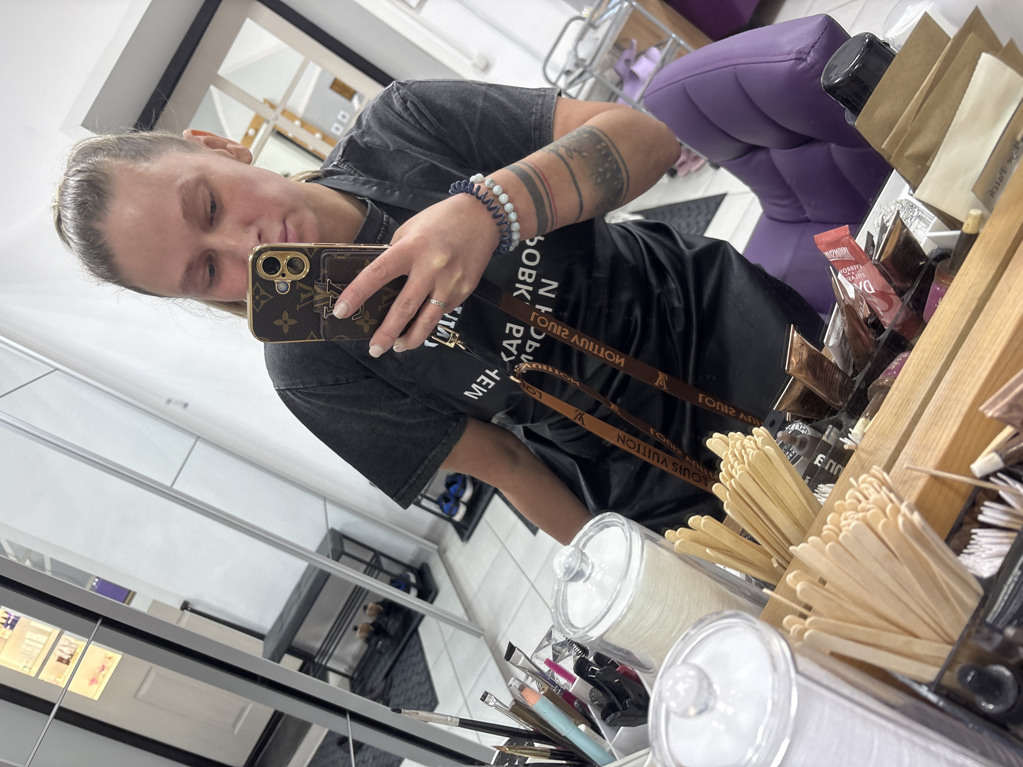 The image is a selfie taken in what appears to be a beauty salon, possibly located in the city of Perm, Russia, given the partial Cyrillic text in the image. A fair-skinned woman with a visible tattoo on her arm is holding up her Louis Vuitton-branded phone. She's wearing a gray-washed t-shirt and two bracelets on her wrist. The salon environment includes makeup brushes, wooden sticks, and various products stored in containers on a wooden shelf. A purple chair is partially visible in the background, along with a reflective surface showing glimpses of other salon equipment.