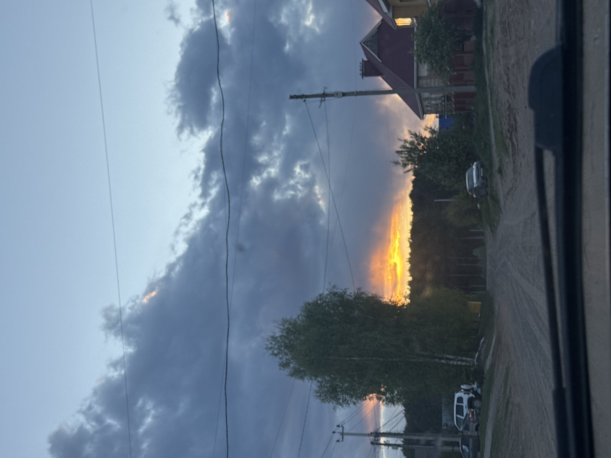 The image shows an early evening scene, potentially a sunset, near Chaikovsky, Russia. The foreground contains the inside of a vehicle, possibly a car. Through the window, the view opens to a rural or suburban street. A dirt road extends into the distance with vegetation along the side. Overhead power lines are visible. A single house with a red roof is partially visible on one side of the road. At least three vehicles are parked along the road. The sky is a mix of blue and gray clouds, with a band of golden light suggesting a sunset. A tree is located to the left.
