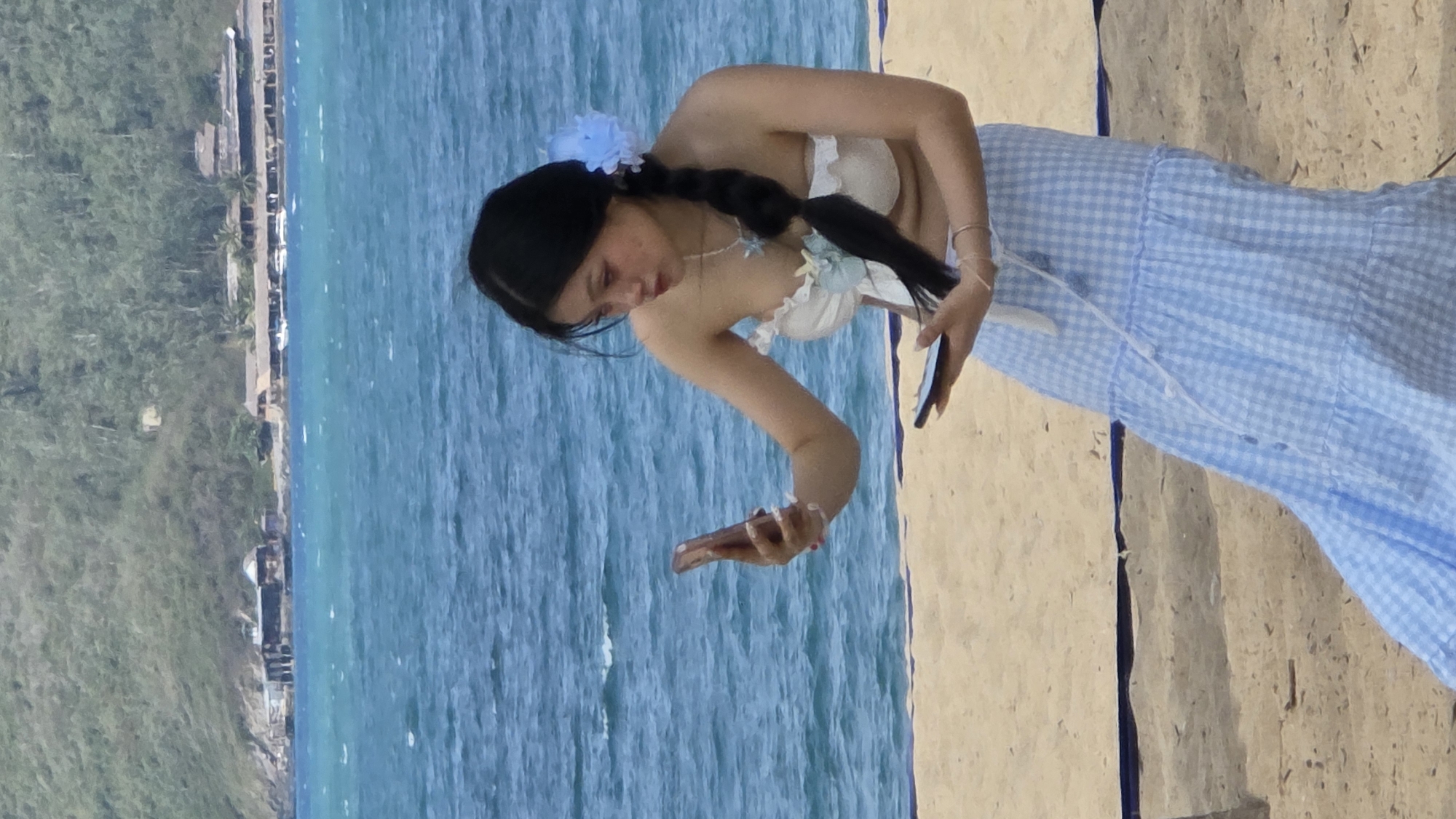 A young woman is photographed on a beach, adjacent to a body of water, likely an ocean. She is wearing a light-colored floral headpiece, a white halter top with floral embellishments, and a blue and white checkered, tiered skirt. She has dark hair in a braid. She is holding a mobile phone in one hand and her other hand is extended out in a gestural pose.
The background includes a body of water that is blue and slightly choppy, with a coastline visible in the distance. The scene includes what appears to be a grassy area and a road, situated behind the beach.
The location is likely in Nha Trang Ward, Vietnam, given the tropical setting.