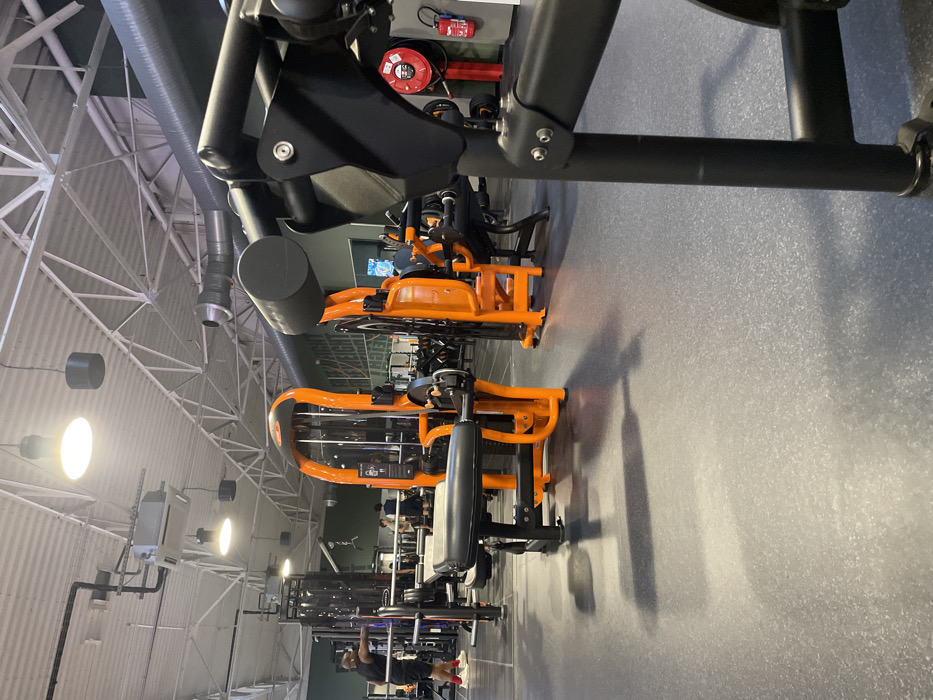 The image depicts the interior of a Basic-Fit gym located at 15 Rue de l'Institut, 14200 Hérouville-Saint-Clair, France. The scene captures a wide view of an exercise area with a dark gray, speckled rubberized floor and an industrial-style ceiling featuring white metal trusses, exposed silver and black ventilation ductwork, and multiple large pendant light fixtures.
In the immediate foreground, a black exercise machine frame with a black padded roller is visible on the right. Centrally positioned is an orange and black leg extension/curl machine with black padded seating. To the left, a male subject, dressed in a black sleeveless shirt, black shorts, and red athletic shoes, stands facing a black barbell rack equipped with a silver barbell. His left arm is extended forward, seemingly in preparation for or completion of an exercise. In the mid-ground and background, several other individuals are present, engaged with various pieces of orange and black exercise equipment, including cable machines and weight benches. The rear wall is a light green/teal color, bearing partial white lettering. On the far right wall, a red fire extinguisher and a red fire hose reel are mounted. The overall environment is brightly illuminated by overhead lighting, casting shadows on the floor.